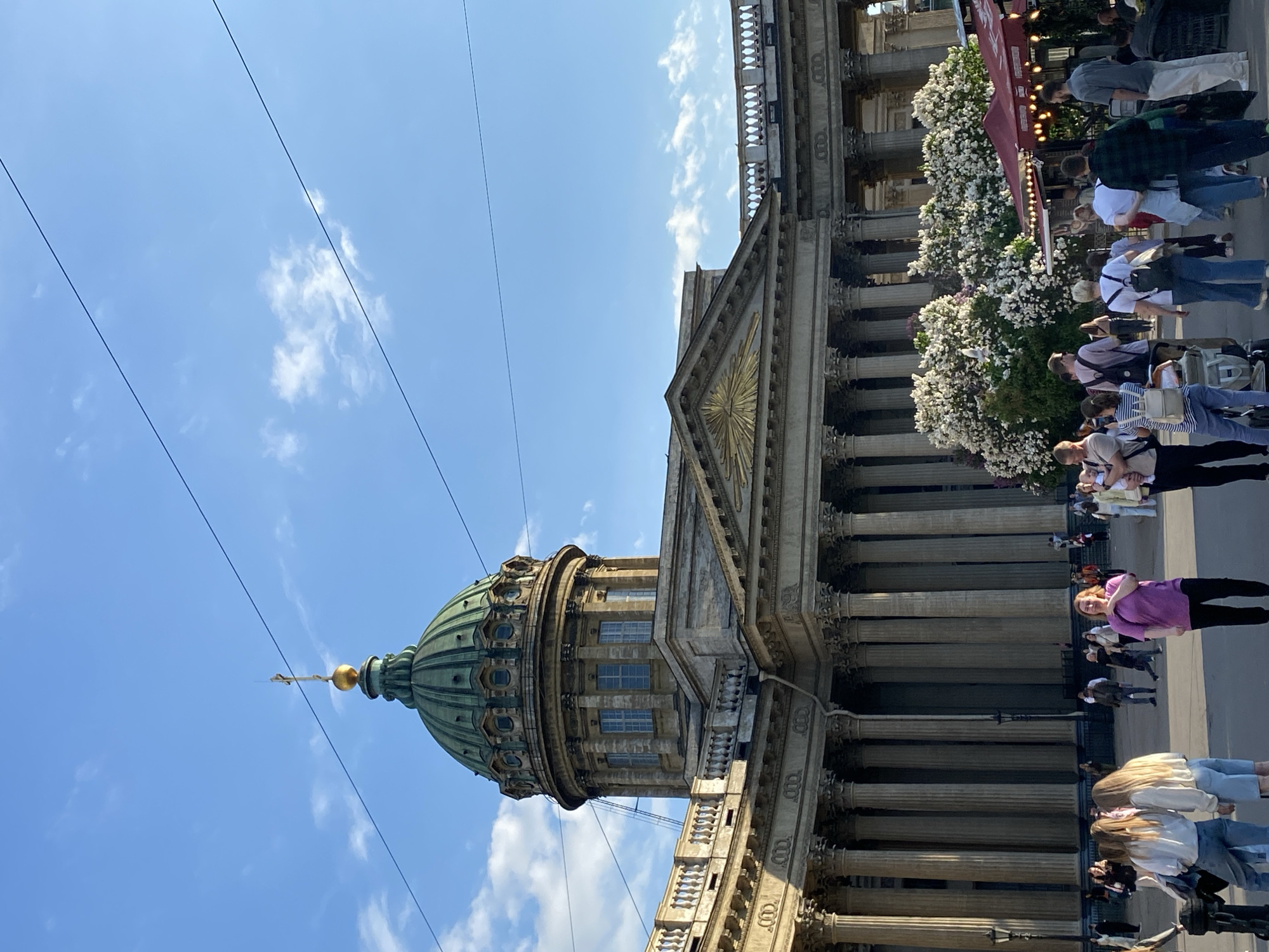 The image, originally rotated, captures a vibrant street scene centered around the grand Kazan Cathedral in Saint Petersburg, Russia, on a bright, sunny day.
The dominant subject is the majestic neoclassical Kazan Cathedral, which occupies the left and center of the frame. Its distinctive green dome, topped with a golden orb and a cross, stands out against the clear blue sky. Below the dome, a multi-story drum with windows is visible. The cathedral's imposing semi-circular colonnade, composed of numerous tall columns, stretches across the right side of the building, featuring intricate architectural details and a pediment with golden sunburst ornamentation.
On the ground level, below the cathedral's colonnade, a bustling crowd of people is visible. Individuals of various ages are seen walking, standing, and interacting, dressed in casual modern clothing. White flowering bushes or trees line the area along the base of the colonnade, adding natural beauty to the urban setting. Partially visible red awnings and sections of text, such as "РЕСТО" (likely part of "ресторан" meaning restaurant), suggest the presence of outdoor cafes or market stalls, indicating general public and commercial activity.
The time of day appears to be mid-day, characterized by strong sunlight and a clear blue sky with a few scattered white clouds. Overhead power lines crisscross the upper portion of the sky. The overall atmosphere is lively and pleasant, with people enjoying the sunny weather around this iconic landmark.