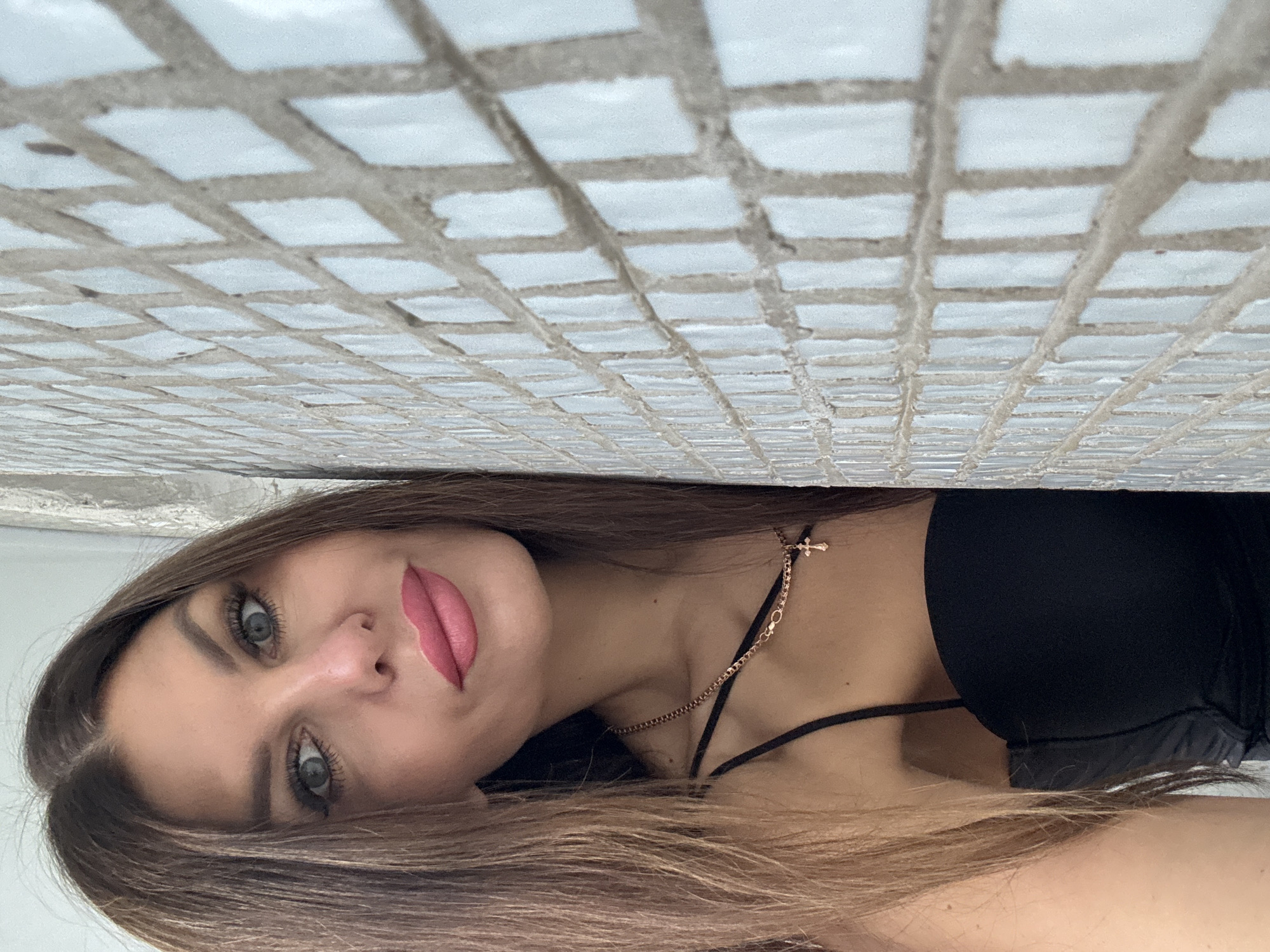 A medium shot portrait shows a woman leaning against a tiled wall. The woman has long, brown hair, blue eyes, and is wearing pink lipstick. She is dressed in a black spaghetti strap top and a gold necklace with a small cross pendant. The tiled wall appears to be a light blue or white with grey grout. The background is out of focus, but appears to be an indoor setting with a light-colored wall. The location is specified as the city of Chaikovsky, Russia.