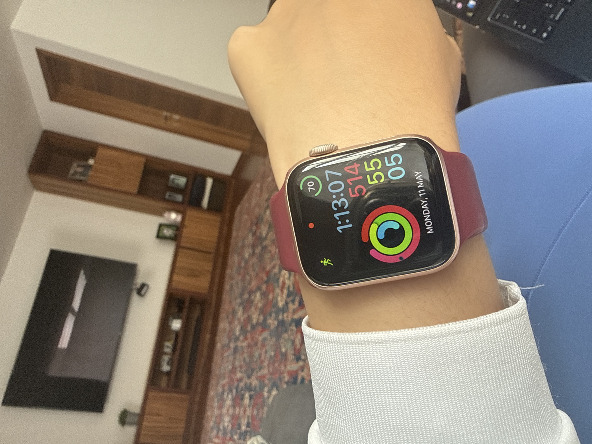 A close-up shot of a person's wrist wearing a rose gold Apple Watch with a burgundy band. The watch face displays the time as 1:13:07, activity rings, and the date, Monday, May 11. The person is wearing a white long-sleeved shirt and a blue garment is also visible. In the background, there's a carpet with a patterned design, a wooden shelving unit, and a black television mounted on the wall. There is no indication of the location being San Bernardino Tlaxcalancingo, Mexico.