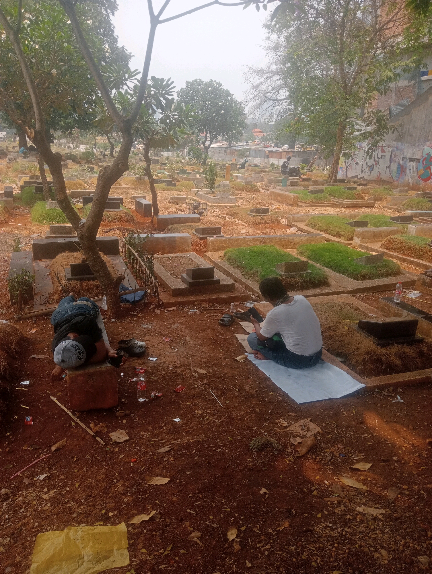 The image depicts a cemetery, likely in South Jakarta, Indonesia, with numerous gravestones and small mounds dotting the landscape. The ground is predominantly reddish-brown soil, with patches of dry grass and scattered leaves. In the foreground, two individuals are present. One person, wearing a white t-shirt and blue jeans, is seated on a light-colored mat, looking down at a mobile phone. Adjacent to them are a pair of dark flip-flops and a plastic water bottle. To the left, another person is lying down on a rectangular stone slab, wearing a black shirt and blue jeans, with a grey baseball cap. A black object, possibly a phone, is in their hand. Scattered on the ground near them are more water bottles, discarded wrappers, and a yellow piece of paper. Trees with sparse foliage are scattered throughout the cemetery, casting shadows and creating a dappled light effect. In the background, more graves are visible, along with hints of urban structures like buildings and what appears to be graffiti on a wall to the right. The overall atmosphere suggests a somber and possibly hot environment.