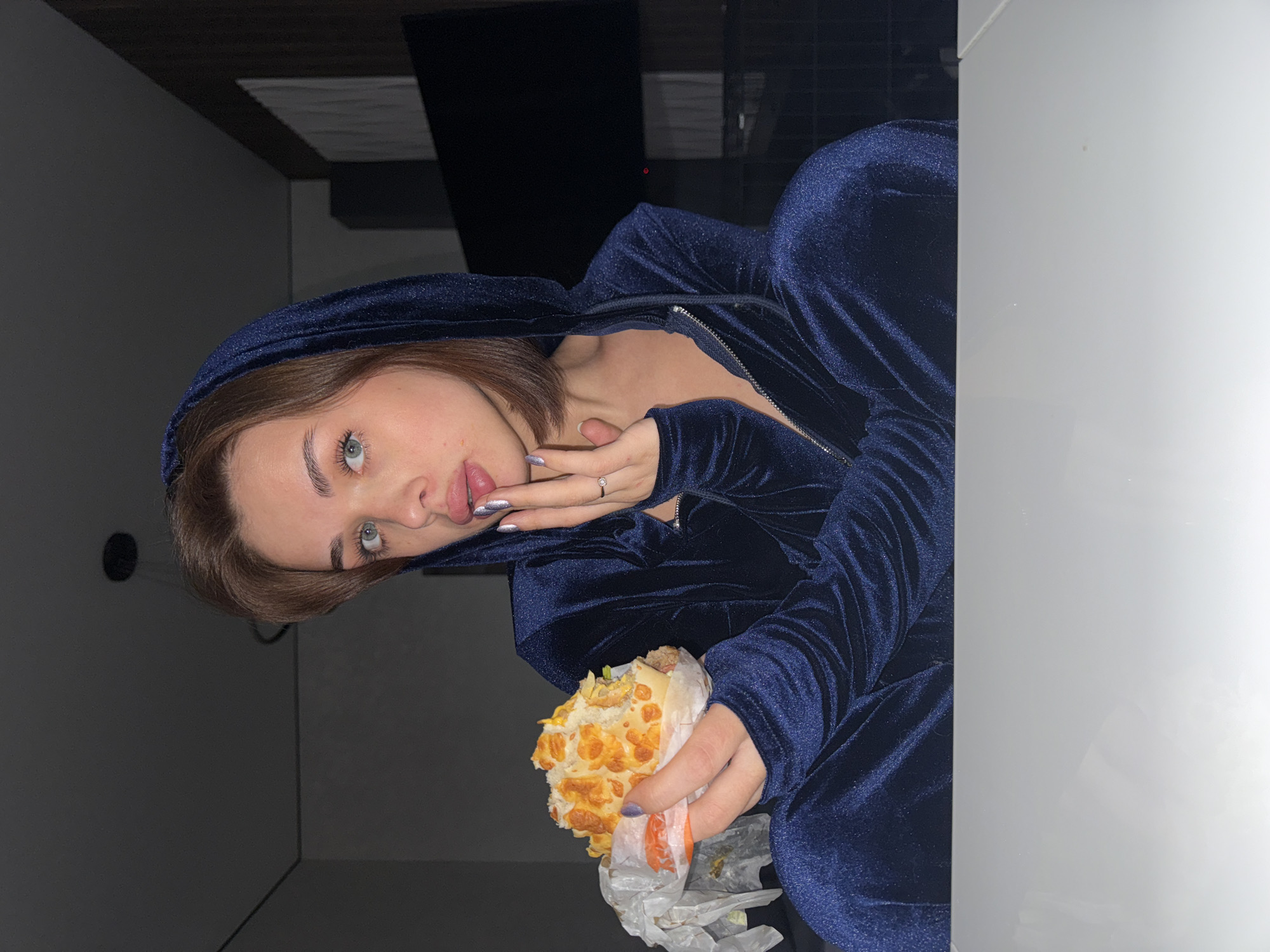 A woman in a dark blue hooded velvet tracksuit is holding a sandwich. She has light brown hair and blue eyes. She is wearing a ring on her left hand. The background is dark, but a ceiling and a wall are visible. No information about the city of Лобаново, Russia can be derived from the image.