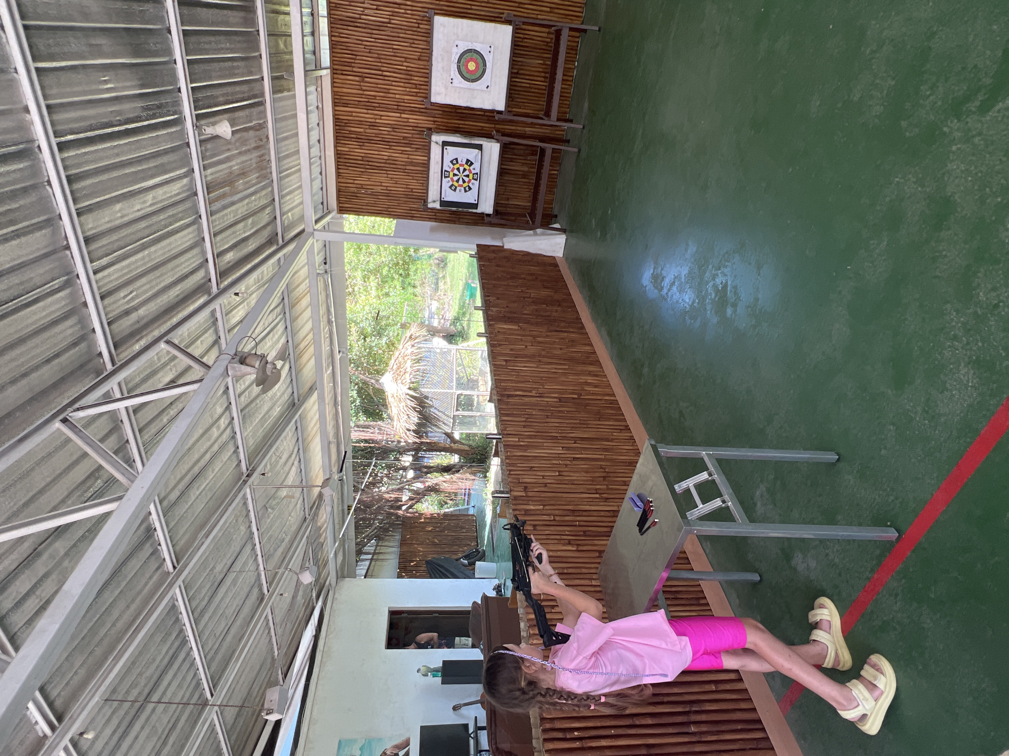 A young girl with long braided hair, wearing a pink shirt and pink shorts, is lying prone on a bamboo floor inside an open-sided structure in Baan Mae Nam, Thailand. She is holding a black air rifle with both hands and appears to be aiming it towards two target boards on the bamboo wall ahead. A metal table with pencils and a notebook rests nearby. The structure has a metal roof, a green floor, and a red line painted on the floor. Outside, foliage and another structure are visible.
