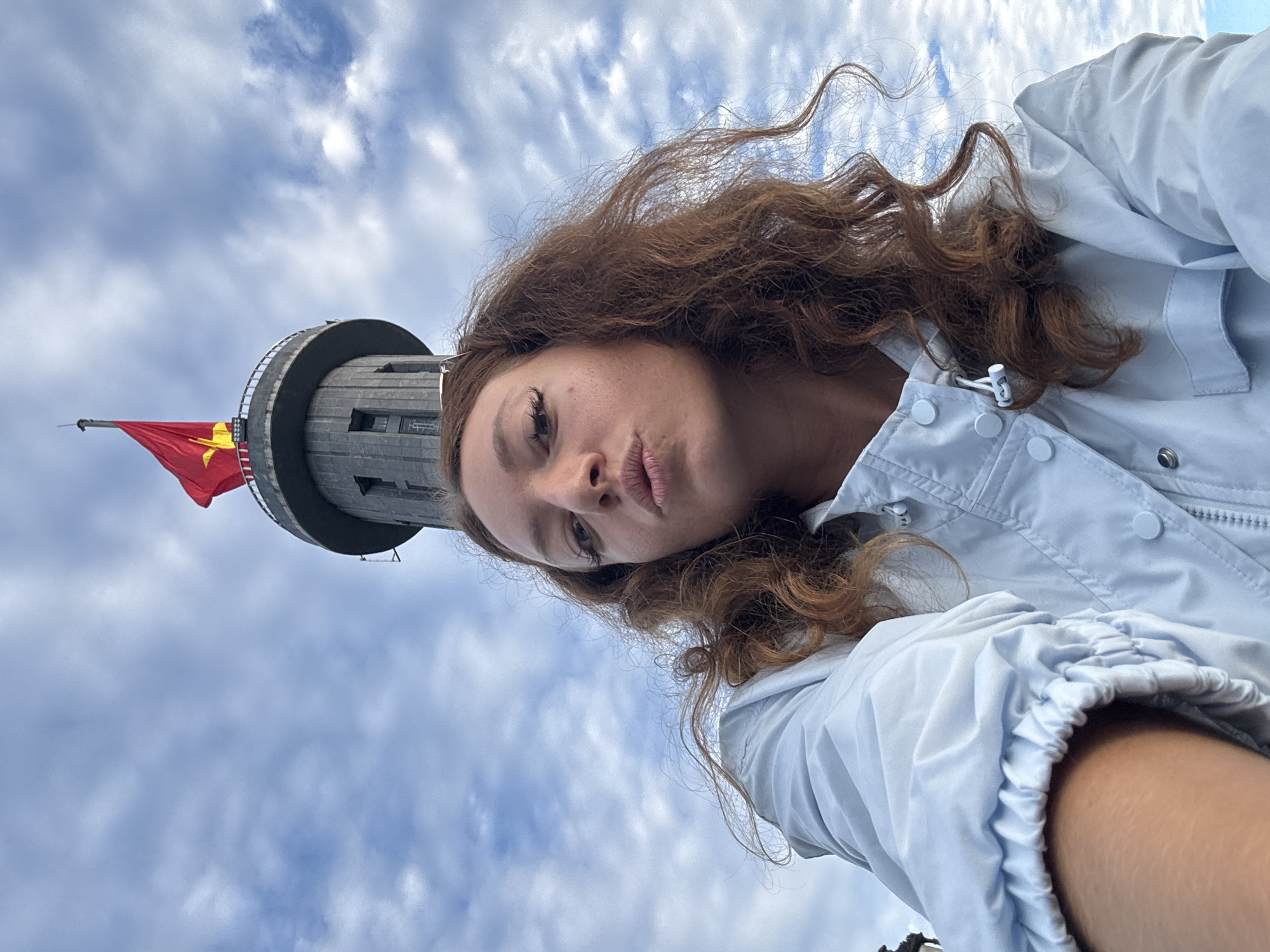 A selfie of a woman with red hair and fair skin, against a backdrop of a tower structure and cloudy sky, taken at the Lung Cu Flag Tower in Lũng Cú Commune, Vietnam. The tower is gray, cylindrical, with multiple levels, and topped with a red flag featuring a yellow star. The woman is wearing a light blue jacket and is positioned so that the tower appears above her head. The sky is filled with white clouds.
