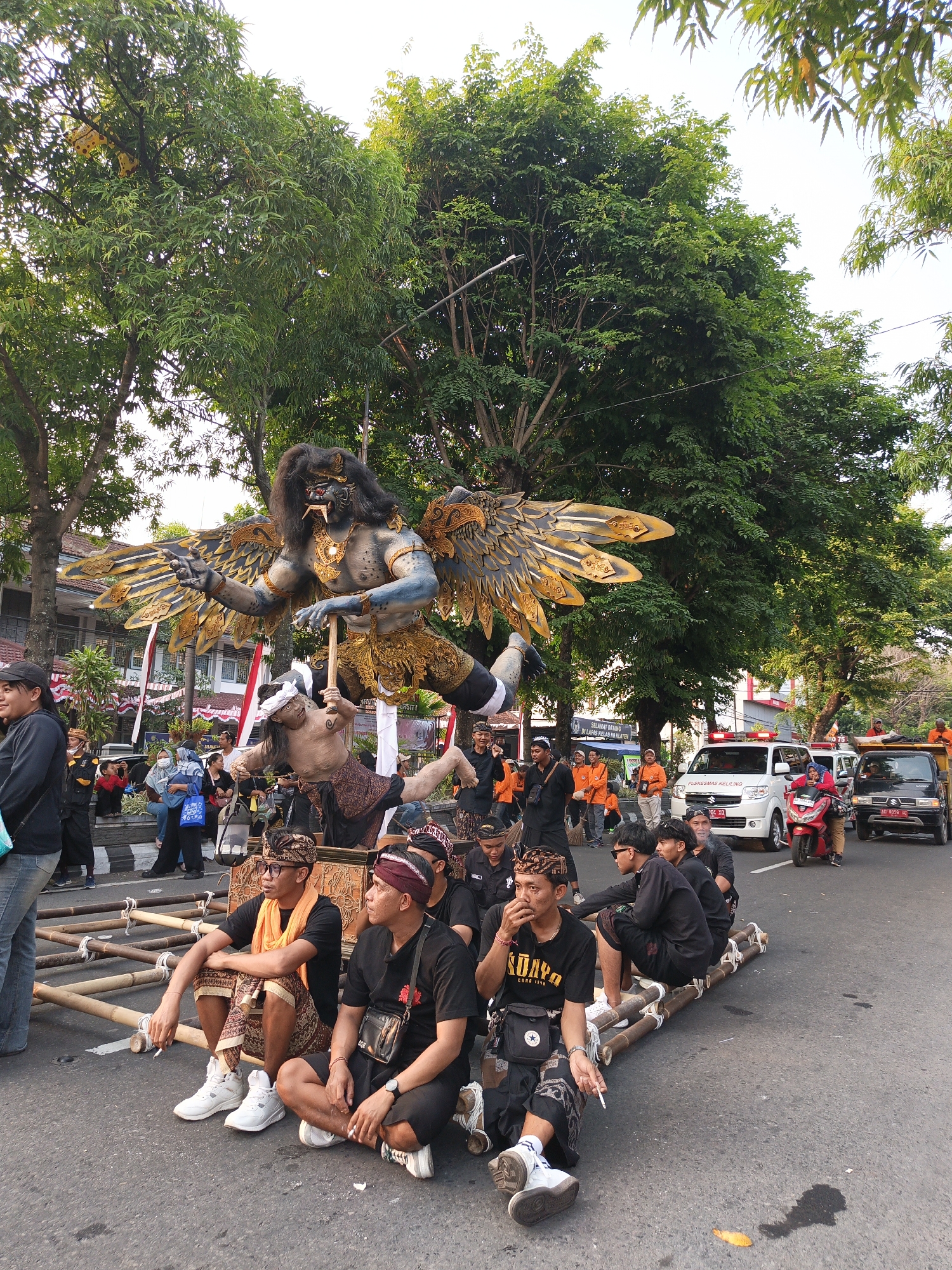 A procession is taking place on a street in Klaten, Indonesia, featuring a large, elaborate effigy of a mythological creature with wings, blue skin, and golden adornments, suspended on a wooden platform carried by several people dressed in traditional Balinese attire. The effigy depicts a winged creature in a dynamic pose, with one hand extended and the other holding a club. Beneath it, another figure, appearing to be human, is shown in a subordinate position. The procession is accompanied by a crowd of onlookers, some of whom are also dressed in traditional clothing. Vehicles, including a white van with red flashing lights, a red scooter, and a black truck, are visible on the street, indicating it is a public event with traffic management. Lush green trees line the street, casting shadows on the pavement. The overall scene suggests a cultural or religious ceremony.
