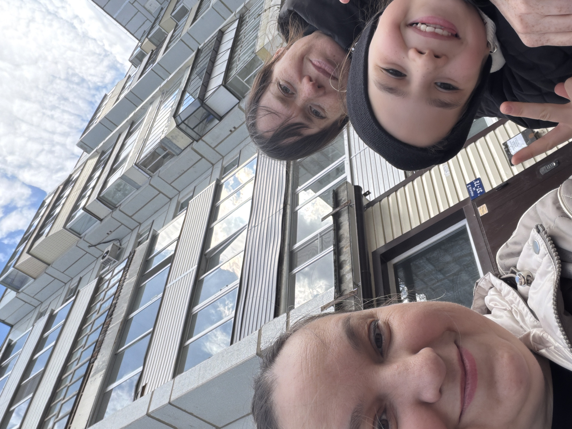 This image is a low-angle selfie featuring three individuals – two adults and one child – taken outdoors against the backdrop of a modern building and a cloudy sky. The perspective is unconventional, with the camera pointed upwards, making the subjects appear to be looking down into the frame.
In the foreground, the top of an adult's head and the side of their face are visible, wearing a light-colored, possibly beige, quilted jacket. Above them, a second adult, with dark hair, looks towards the camera with a gentle smile, dressed in a black jacket. Next to this adult, a child, likely a girl, smiles widely at the camera, wearing a black beanie and a black quilted jacket. Her right hand is raised, making a peace sign gesture.
The background is dominated by a multi-story building, characterized by numerous large, rectangular windows that reflect the bright, cloudy sky. Portions of the building feature vertical paneling, and an external air conditioning unit is visible on an upper level. The bright, diffuse light suggests it is daytime.
A small blue plaque with white Cyrillic text is visible on a surface behind the child. The text reads "Проспект 2" and "37-12," which translates to "Avenue 2" and a number, likely an address or building identifier. The overall scene captures a candid moment, with expressions ranging from neutral to happy, set in what appears to be an urban environment in Chaikovsky, Russia.