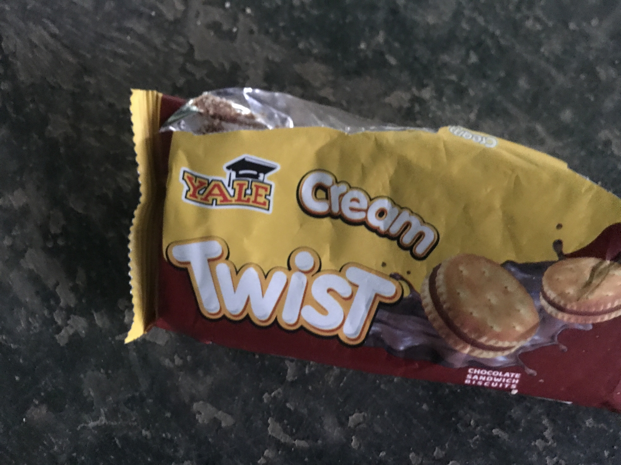 A product packaging of "Twist Cream" chocolate sandwich biscuits is displayed on a dark, textured surface that could be asphalt or concrete. The packaging is mostly yellow with red accents. The word "Twist" is prominently displayed in large white, orange-bordered letters vertically on the left side of the package. Below it, also vertically oriented on the red portion of the packaging, are the words "CHOCOLATE SANDWICH BISCUITS" in white capital letters. The word "Cream" is written vertically on the yellow part of the packaging in white, orange-bordered letters. A logo featuring an open book with a graduation cap is visible on the upper right side of the yellow section. Two chocolate sandwich biscuits are partially visible on the lower left of the packaging, with chocolate filling oozing out. The lighting is dim, suggesting an outdoor environment, potentially on a street or ground surface in Jos, Nigeria. No people are visible in the image.