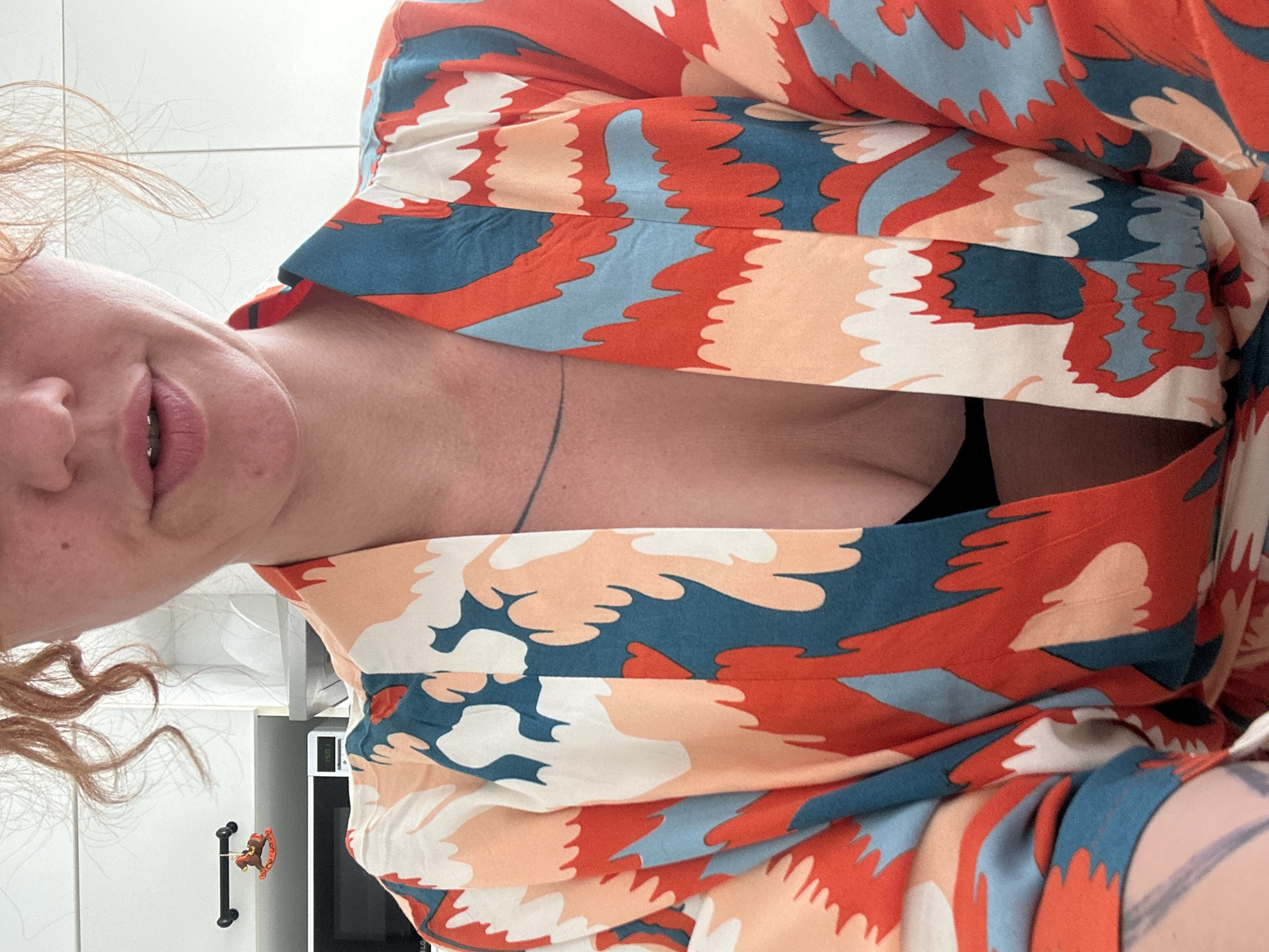 A person is posing indoors, wearing a colorful top with a pattern of red, orange, blue, and white, possibly in a kitchen setting. The person has fair skin, reddish-blonde curly hair, and visible tattoos on their arm. A cabinet with a decorative handle is visible in the background, along with a microwave. The context of Sochi, Russia, cannot be confirmed based on the image.