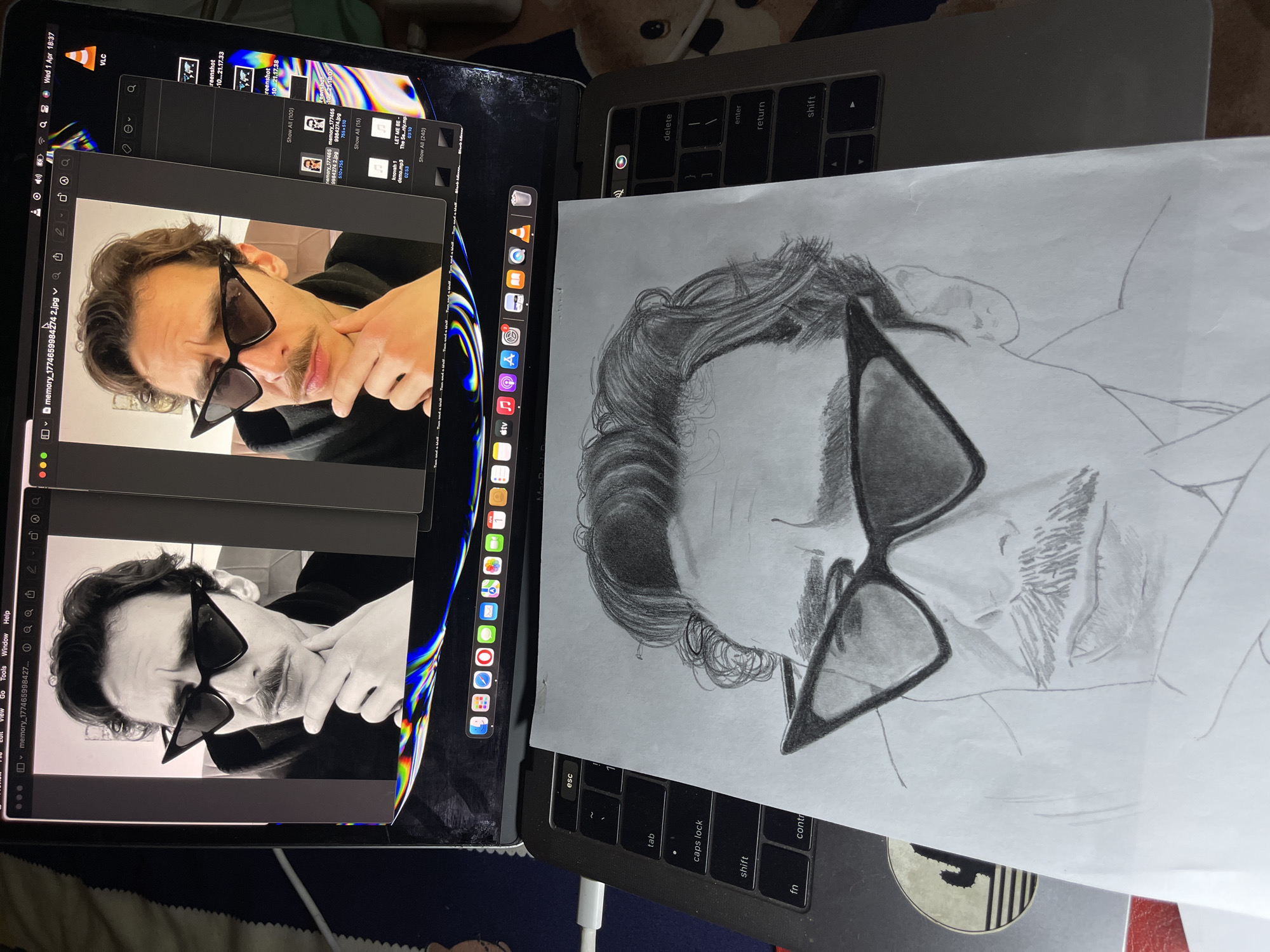 The scene depicts a drawing of a man wearing sunglasses next to a laptop displaying reference images of the same man. The drawing is on a white sheet of paper, and it's placed directly on top of the laptop's keyboard. The laptop screen shows image files and a video player on the left and icons to the right. The subject of the image is located on a gray desk and the color palette includes grayscale, white, black, and some color from the application icons. The location is not possible to determine.
