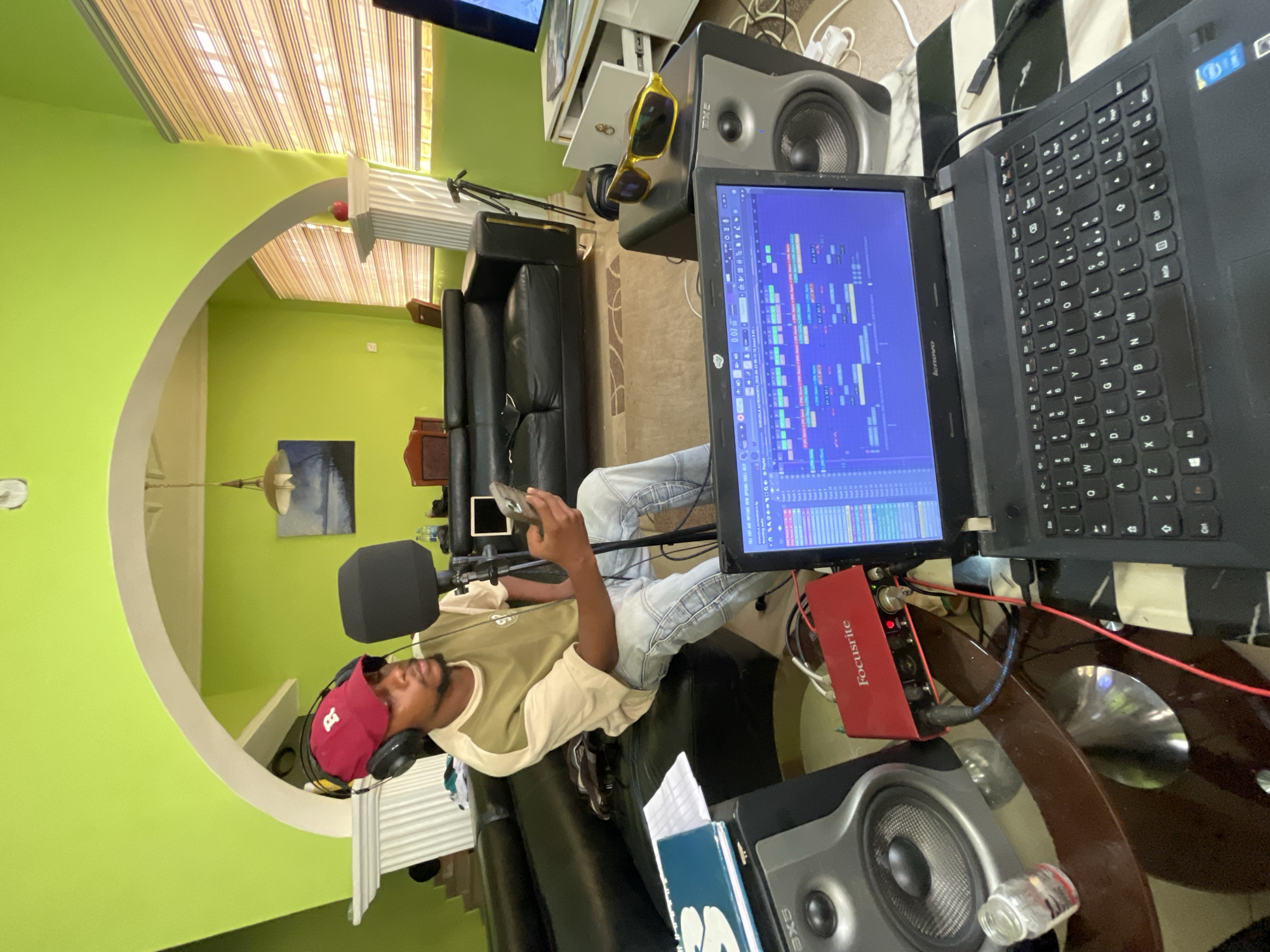 The scene depicts an individual, likely a young man, in what appears to be a home recording studio setup. He is wearing a red cap and headphones, and is seated on a black leather couch. He holds a mobile phone in his hands, possibly interacting with it. A microphone with a pop filter is positioned in front of him, suggesting he is involved in recording or voice work.
The environment includes a laptop displaying audio editing software, a Focusrite audio interface, and studio monitors (speakers). A table supports the equipment. The background reveals a living room setting with a green wall, an arched doorway, a painting, and window blinds.
While the image lacks specific details to pinpoint a precise location within Abuja, Nigeria, the setup and domestic environment suggest a home-based recording activity within the city.