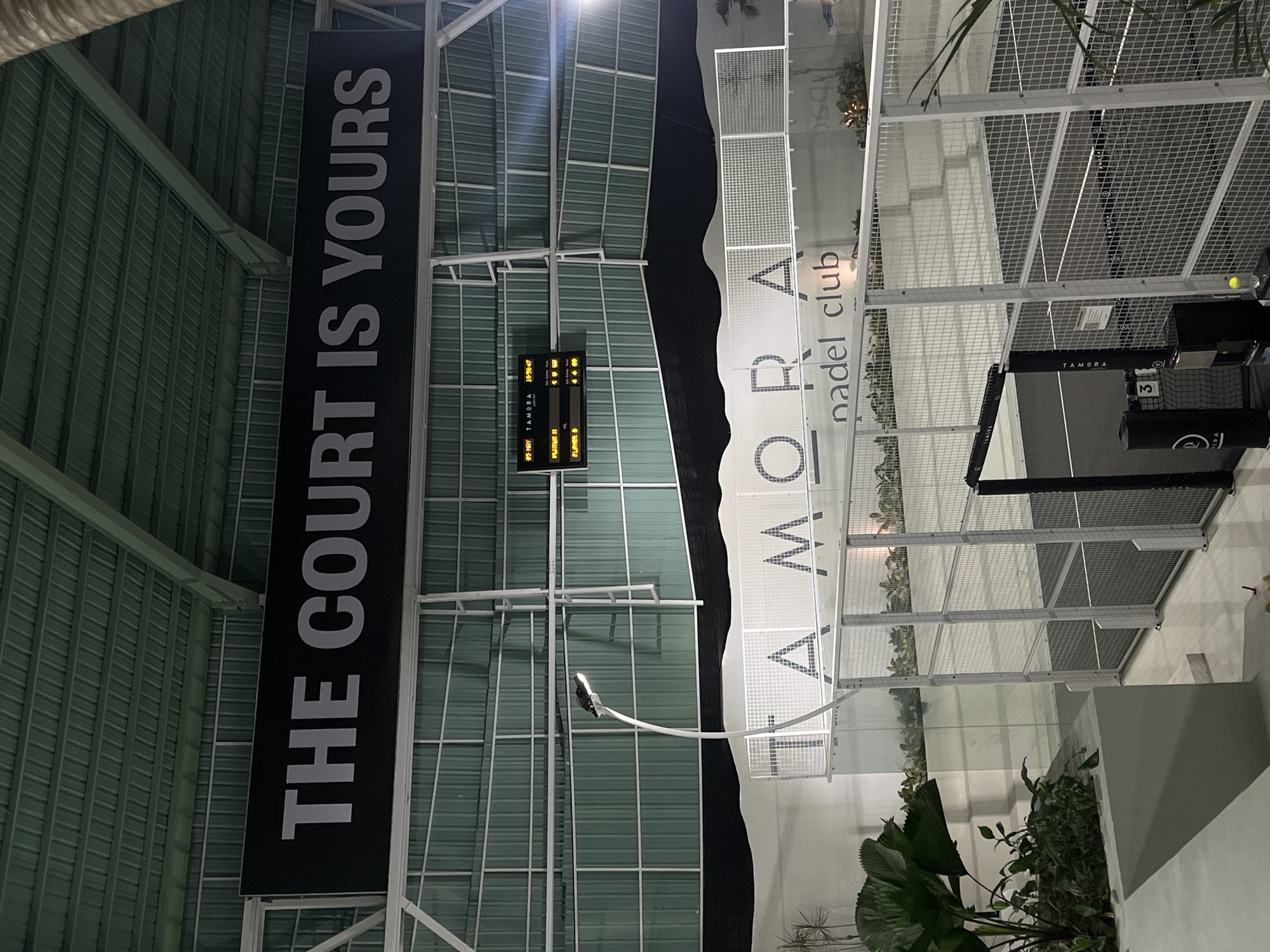 This is an indoor or covered padel tennis club, likely the TAMORA nadel club based on the large sign. The scene is illuminated by overhead lighting. A scoreboard displays game information. Banners with text 'THE COURT IS YOURS' are visible. The location is North Kuta, Indonesia, as indicated on the scoreboard. Various metallic structures make up the building's framework and netting around the court. Potted plants are located around the court. A black net and a device with the number 3 on it are positioned beside the court.