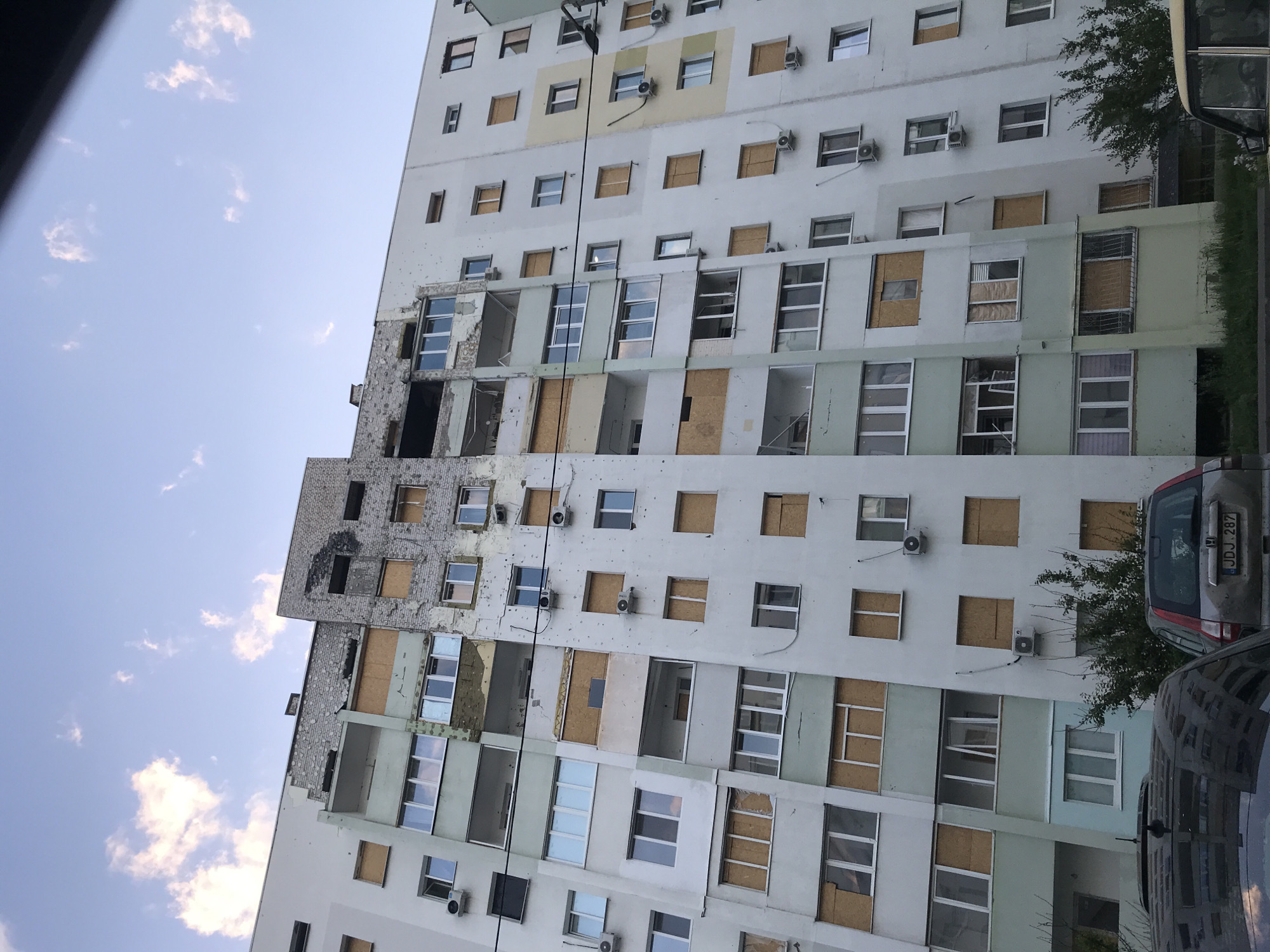 A low-angle shot shows a multi-story building with damage, likely from shelling or explosions. The building's facade is marked with impact holes and areas of destruction. Some windows are boarded up with plywood, while others are shattered or missing entirely. The building's structure is a mix of white, gray, and light green colors. Air conditioning units are mounted on some of the undamaged sections of the building. A light blue sky with scattered clouds is visible in the upper left of the frame. Parts of vehicles are visible in the foreground. The license plate on one of the cars reads "JDJ 282." The building does not appear to be in Lima, Peru, due to the European-style architecture and license plates on the cars. The location is likely in a war-torn area, possibly in Ukraine or a similar conflict zone. There are no visible people in the image.