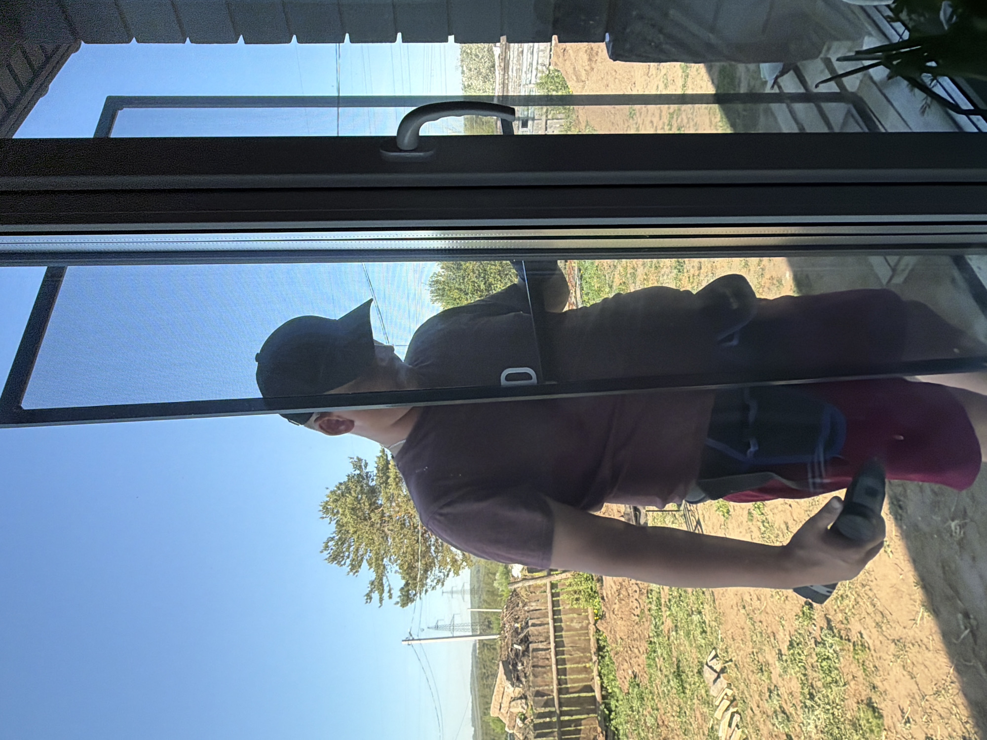 The image appears to be taken from inside a building, looking out through a window. A person is reflected in the window, wearing a dark baseball cap, a maroon t-shirt, and red shorts. The person is holding a black object in their hand. Outside the window, the background shows a clear blue sky, trees, a field of green grass, a fence, and power lines. The building appears to be located in a rural area with vegetation.
It is difficult to specify exact location in Chaikovsky, Russia without more details.