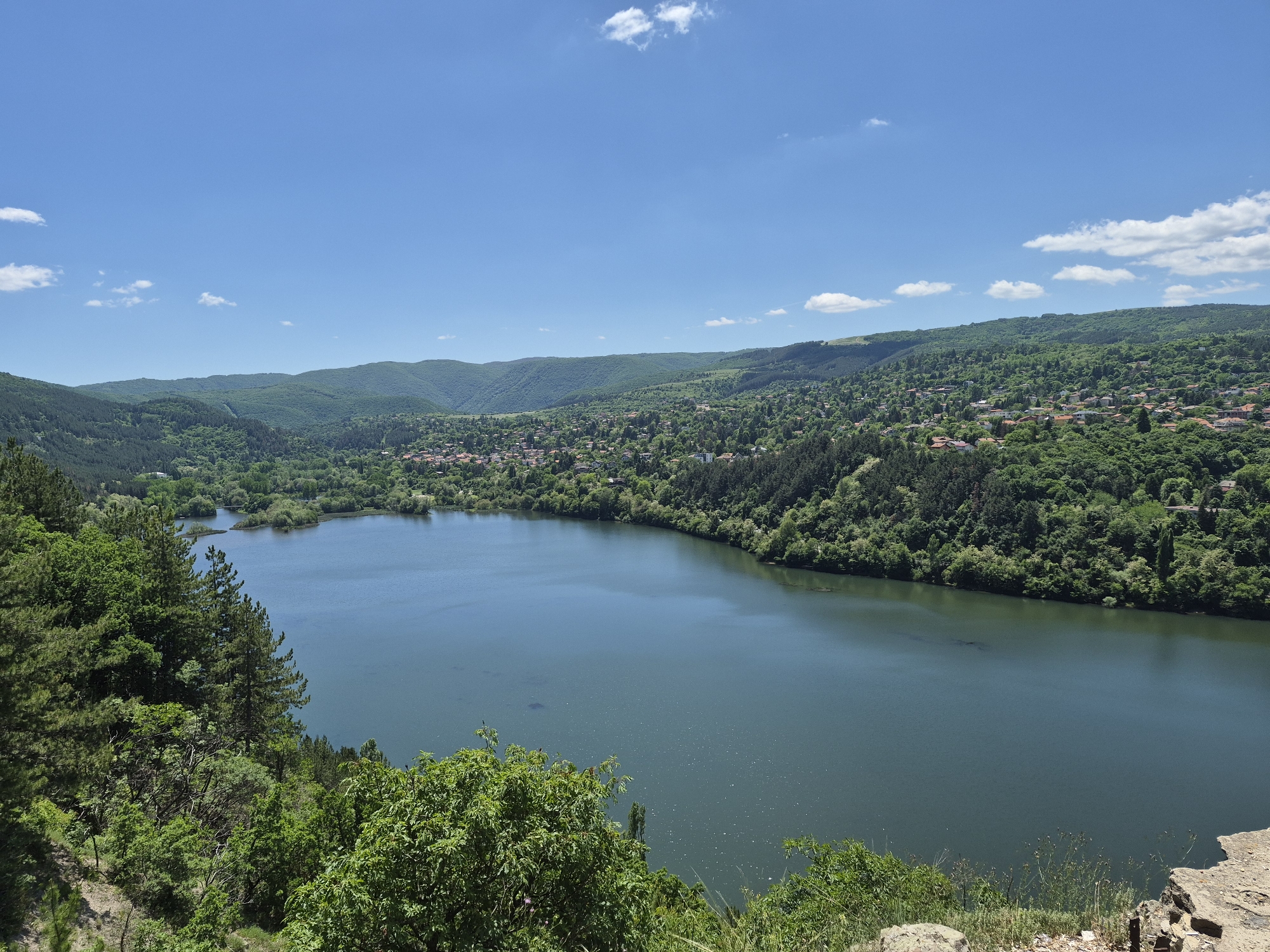 The image presents a wide-angle, daytime view of a lake surrounded by lush, green hills and a small settlement. The sky is blue with scattered clouds. The water appears still, reflecting the sky and surrounding landscape. Along the lake's edges, dense forest vegetation transitions into patches of residential housing in the background. The foreground is comprised of trees and shrubbery, suggesting the viewpoint is from an elevated position. The location is in the city of Unknown, Unknown.