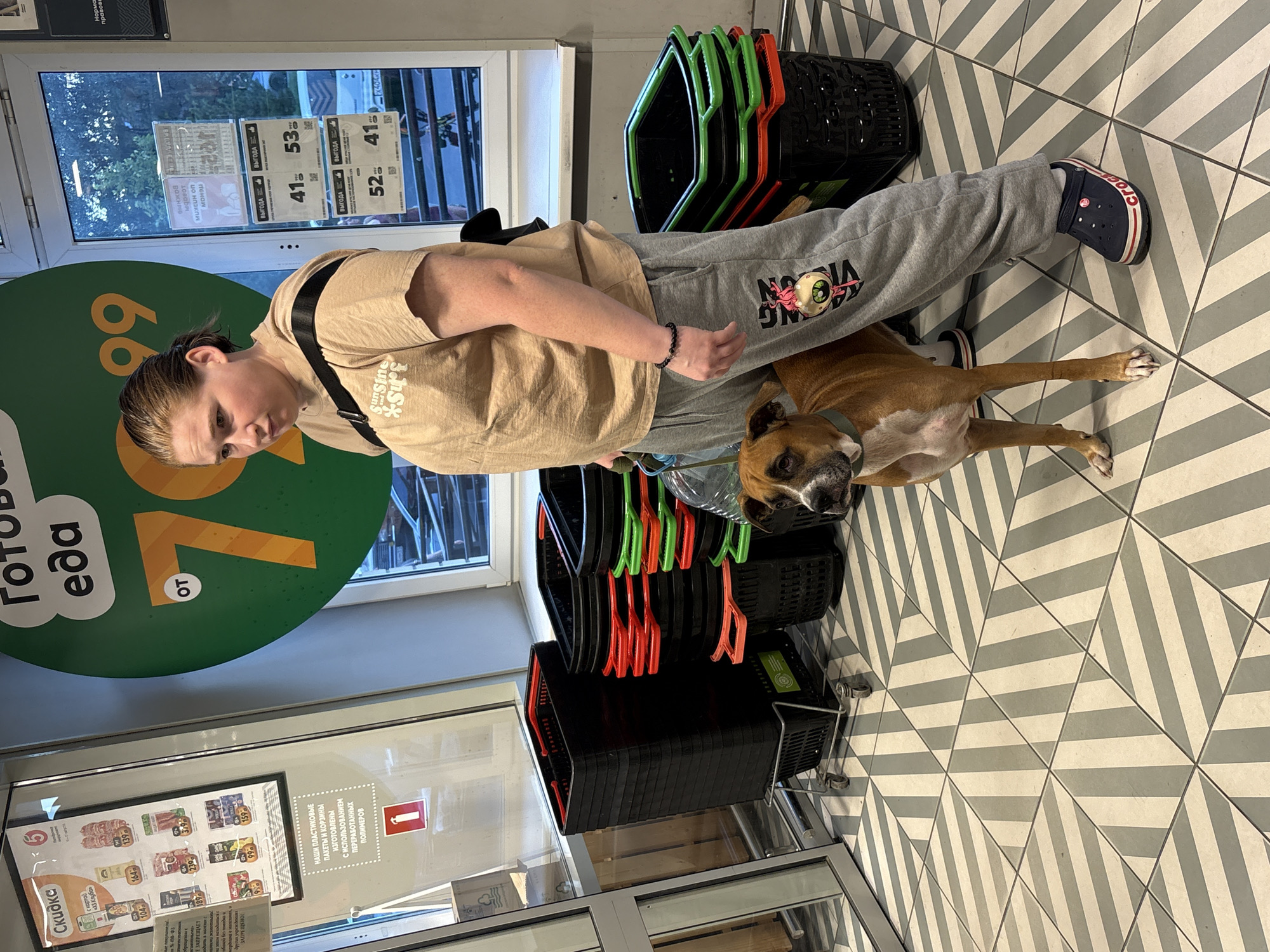 A young adult woman, with light brown hair pulled back, wearing a beige t-shirt and gray sweatpants, stands in a supermarket in Rostov-on-Don, Russia. She is accompanied by a medium-sized, tan-colored dog on a leash. The woman is looking down and to her left. The dog is looking forward.
The environment is a brightly lit supermarket with a patterned floor consisting of light gray and beige diagonal stripes. In the background, there are stacks of black and colored shopping baskets, as well as a large green circular sign with the text "Готовая еда" (Ready food) and the number "70%" prominently displayed. Posters and advertisements are visible on glass windows in the background. The overall scene suggests a shopping trip.