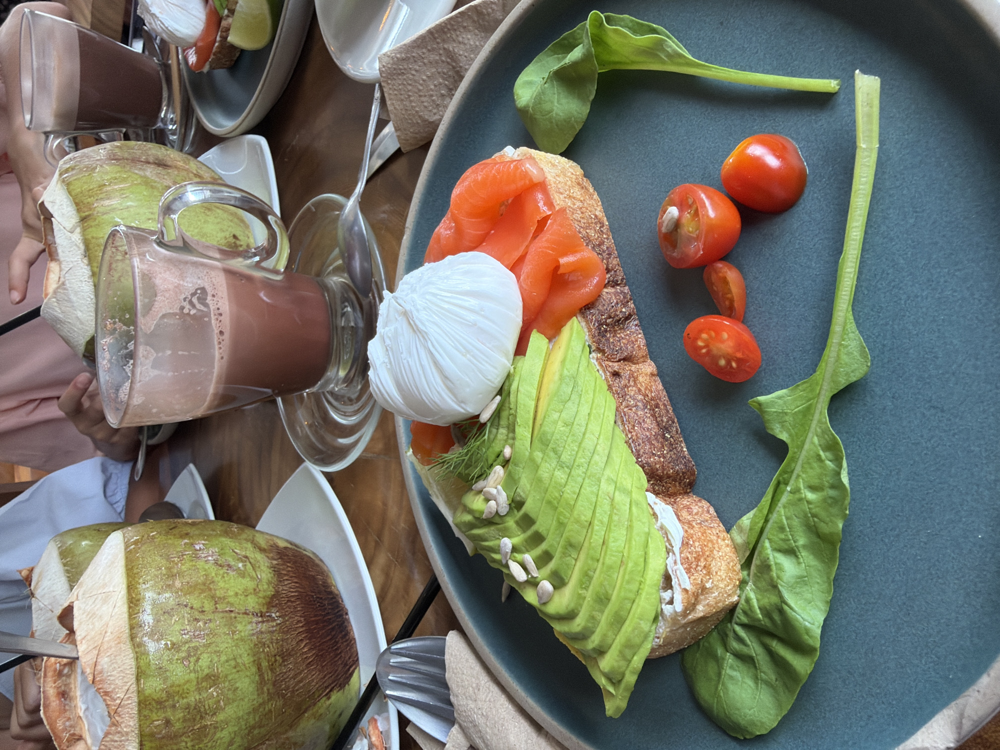 In a close-up, overhead shot, a rustic wooden table serves as the backdrop for a vibrant brunch spread in Ko Samui District, Thailand. Two whole coconuts, their green husks partially peeled to reveal the white interior and a straw inserted into one, sit on white plates, suggesting a refreshing beverage. Nearby, a glass mug filled with a frothy, chocolate-colored drink rests on a saucer, steam hinting at its warmth.
The main focus is a delectable open-faced sandwich on a dark, crusty bread. The toast is generously topped with cream cheese, thinly sliced smoked salmon, and fanned avocado slices. A perfectly poached egg, its white fluffy and yolk intact, sits atop the avocado. Scattered across the avocado are sunflower seeds, adding texture. The sandwich is garnished with a sprig of dill and accompanied by a few halved cherry tomatoes and a vibrant green leaf of spinach or arugula. The entire dish is presented on a deep teal plate, creating a visually appealing contrast. The scene captures a moment of leisurely dining, likely at a cafe or restaurant, given the presentation and the presence of other diners subtly visible in the background.