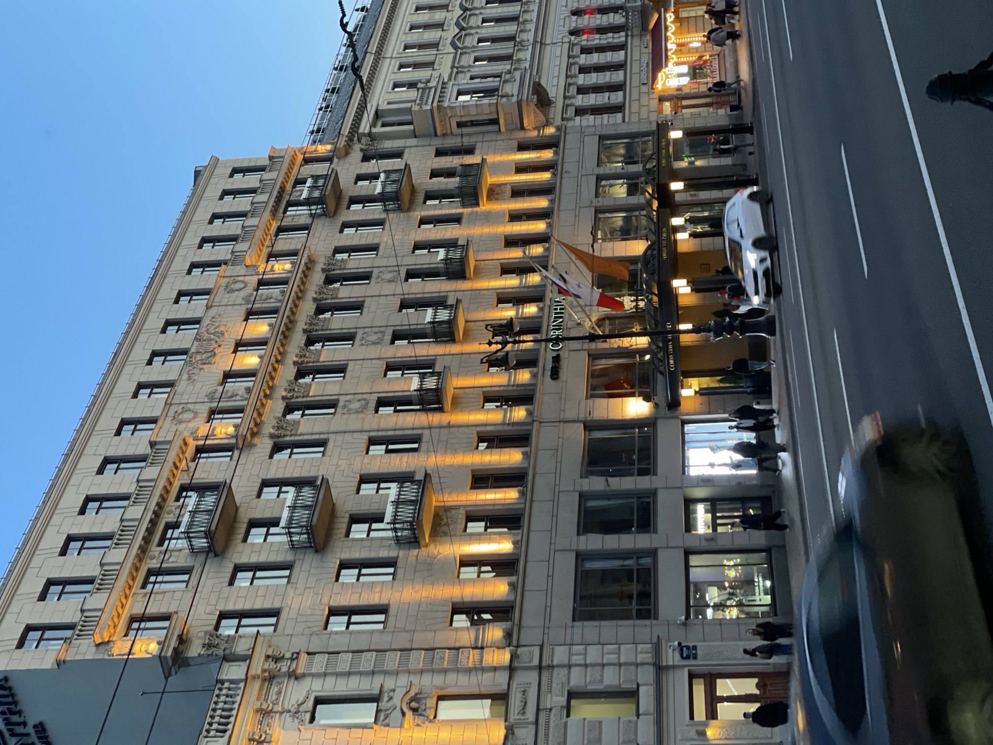 The image captures a grand, multi-story building at dusk or early evening, likely in Saint Petersburg, Russia, as suggested by the location context. The sky is a clear, deep blue, indicating twilight, while numerous warm yellow lights illuminate the building's facade and interior spaces, creating a striking contrast.
The central subject is a majestic architectural structure, possibly a hotel, characterized by its classical or neo-classical design, featuring intricate decorative elements, ornate cornices, and numerous rectangular windows. Several windows and architectural details are highlighted by external lights. Flags are displayed near the main entrance, with one clearly showing the white, blue, and red horizontal stripes of the Russian flag, and another, possibly a hotel or corporate flag. Text "CORINTHIA" is prominently visible on the building's facade, identifying it as the Corinthia Hotel.
Below the building, a city street is visible, with dark asphalt and clear white lane markings. Several pedestrians are walking on the sidewalk adjacent to the building. A white car is seen moving along the street, and another vehicle is partially visible in the foreground on the right. Illuminated storefronts and signs, some reading "THE GRILL", line the ground floor of the building, suggesting commercial establishments. The overall atmosphere is calm, depicting a typical urban scene during the evening transition from day to night.