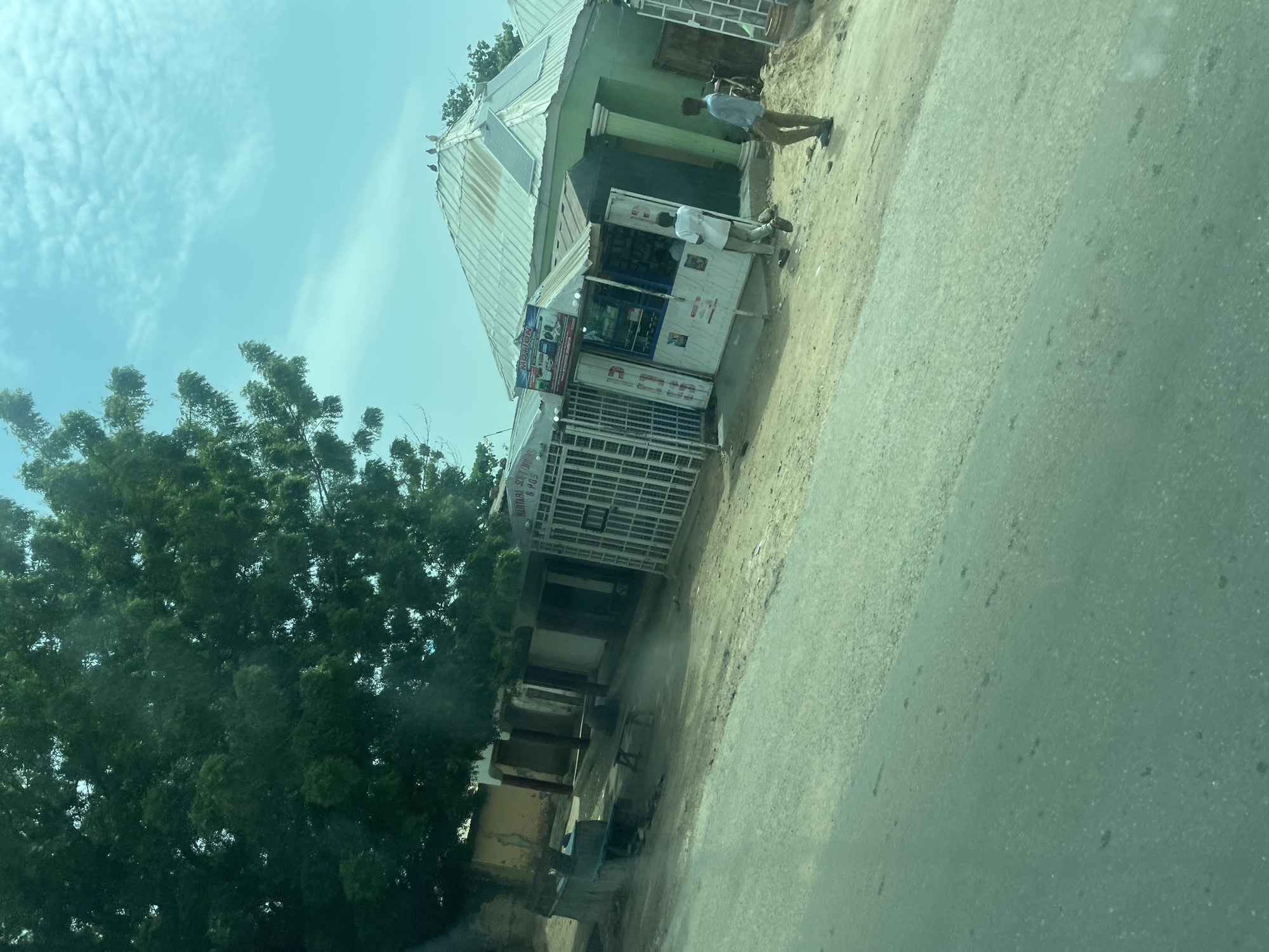 A street view shows a storefront on the right side, featuring a green building with a corrugated metal roof. Two solar panels are visible on the roof. In front of the green building is a smaller, white stall with a metal security gate. A sign on the stall reads "MOTION." Two men are standing near the stall. One man, wearing a white shirt and dark pants, is walking away from the stall. Another man, in a blue shirt and khaki pants, is standing and looking to the right. A large tree with dense green foliage dominates the left side of the image, casting shadows on the ground. Several other buildings are visible further back, some with what appear to be arches. The ground is dusty and unpaved. A paved road occupies the foreground. The sky is partly cloudy. This scene is consistent with a street in Chabal, Nigeria, a location known for its bustling local markets and small businesses.