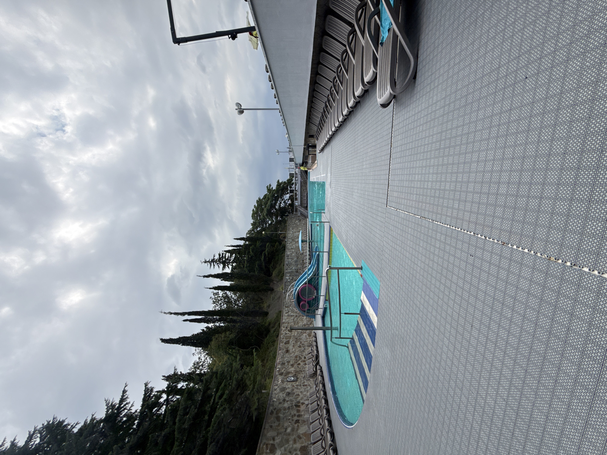 An outdoor swimming pool area is pictured under a cloudy sky in Массандровский поселковый совет, Ukraine. The foreground features a grey, tiled deck with a repeating geometric pattern, leading to a turquoise swimming pool with a vibrant blue and white tiled step area. A blue and pink waterslide is visible in the pool, with a small umbrella nearby. To the left of the pool, a stone wall separates the area from a hillside covered in dense green trees, including several tall cypress trees. A row of empty lounge chairs is arranged along the edge of the deck, facing the pool and the hillside. To the right, more lounge chairs are lined up next to a white building with a dark railing. The sky is overcast with scattered clouds, casting a diffused light over the scene. There are no people visible in the image.