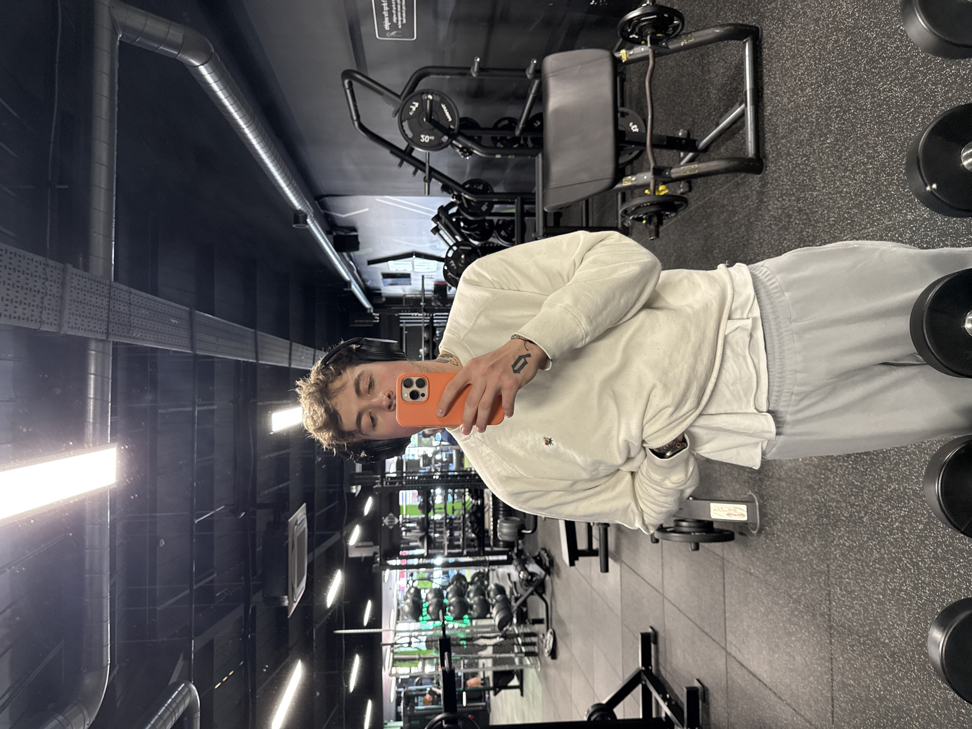 This media file features a selfie taken by a young individual inside a gym located in Letchworth, United Kingdom.
The primary subject is a person with short, curly brown hair, wearing black over-ear headphones. They are dressed in a cream or off-white long-sleeved sweatshirt, possibly made of a soft material like velour, and light grey sweatpants. A white undershirt is visible beneath the sweatshirt. The person holds an orange-cased smartphone in their right hand, using it to capture the image. A tattoo resembling a stylized letter (possibly a Gothic 'D') is visible on their right hand/wrist area.
The setting is a well-lit modern gym. The floor is dark grey with a speckled, rubberized texture typical of fitness facilities. The ceiling and upper walls are dark. Bright, rectangular fluorescent light fixtures provide illumination, casting reflections. Industrial-style silver ventilation ducts and cable trays are visible along the ceiling.
In the background, various pieces of gym equipment are present. To the right of the person, a black weight machine with a padded seat is prominent, featuring a weight plate clearly marked with "50". Further into the gym, rows of dumbbells or weight plates are stacked vertically, and other exercise machines can be observed, indicating a comprehensive fitness environment. The indoor lighting suggests the photo was taken during operational hours, but the specific time of day is not discernible.
A small, unreadable sign is visible on the wall in the upper left background (appearing upside down in the original image), which might contain gym rules or warnings.