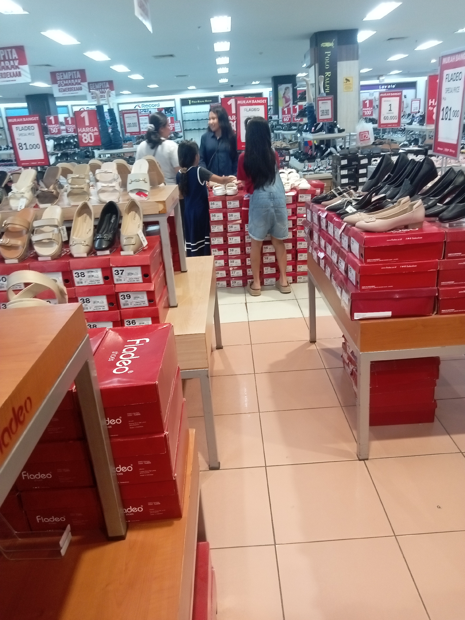 The image depicts the interior of a shoe store in Batam, Indonesia, likely a department store, filled with various shoe displays and shoppers. Prominent signage indicates sale promotions with prices such as "81,000" and "181,000". Various styles of women's shoes, including sandals and closed-toe heels, are displayed on shelves and in boxes. One woman, wearing a dark blue shirt and dark pants, is interacting with a younger girl in a denim overall skirt, who is facing away from the camera. Another woman with dark hair pulled back is visible in the background, also appearing to be a shopper or employee. The store environment features tiled flooring and bright overhead lighting.