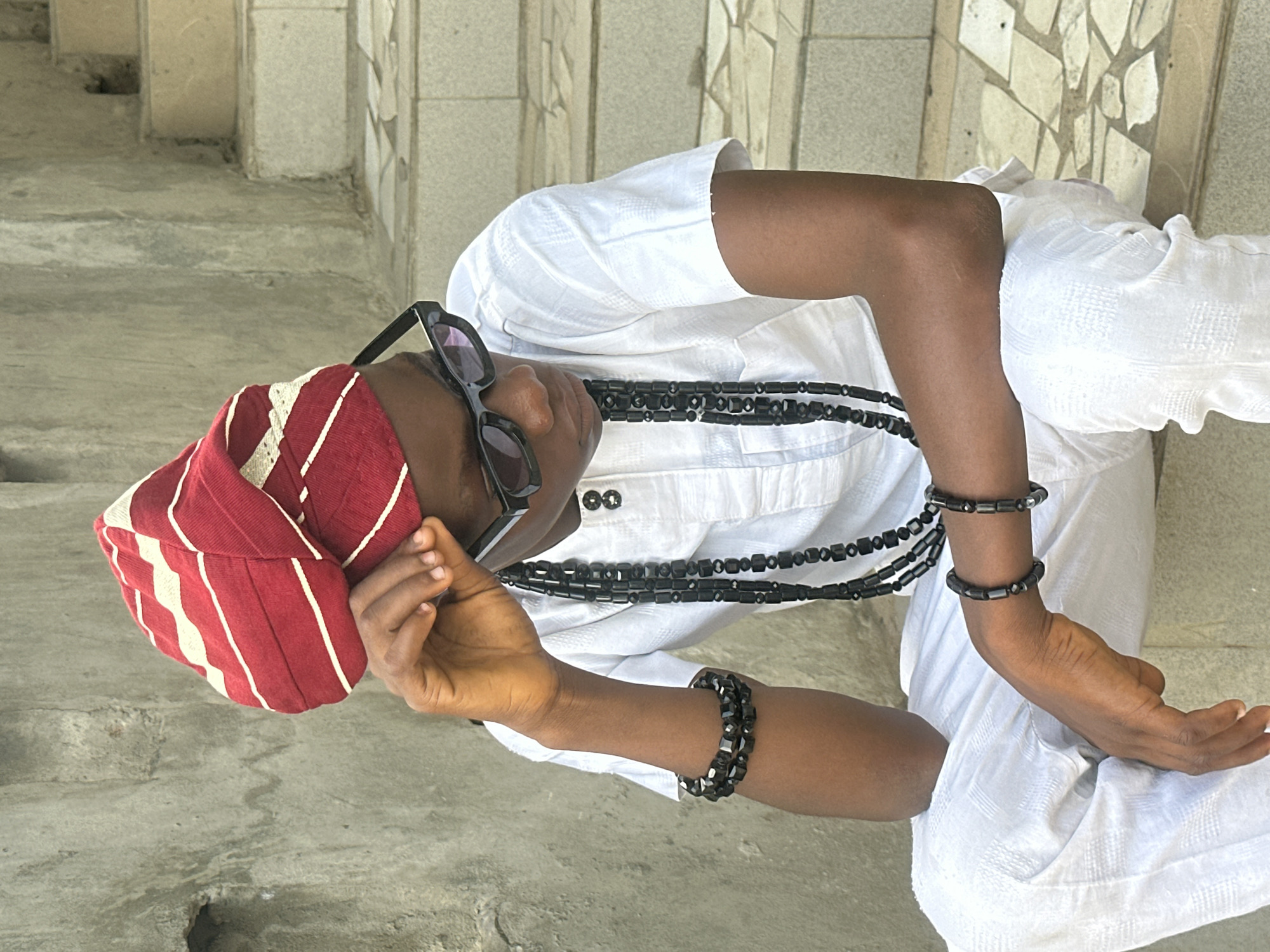 A young Black male is sitting on stone steps in Badagry, Nigeria. He is wearing a white traditional outfit consisting of a tunic and trousers. On his head, he has a red and white striped turban. He is also wearing black sunglasses and multiple strands of black beaded necklaces and bracelets. His left hand is resting on his knee, while his right hand is holding the edge of his turban. The background consists of a textured concrete wall and a set of stone steps.