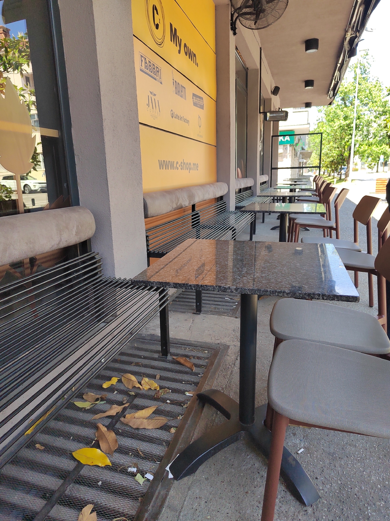 This image captures the outdoor seating area of a cafe or shop, likely located in Podgorica, Montenegro, given the context. The scene is devoid of people, suggesting it might be early morning, off-peak hours, or closed.
The setting features a row of tables and chairs extending along the building's exterior. The building itself has a light gray facade, and a prominent yellow panel displaying brand names and a website. Overhanging the seating area is an awning with ceiling lights and a mounted industrial-style fan, along with a wall-mounted heater, indicating preparations for various weather conditions.
The tables are rectangular with a dark, speckled, granite-like surface and black metal bases. The chairs are modern, featuring light gray fabric seats and backs, supported by brown wooden frames and black metal legs. Along the building wall, there are built-in benches with metal slat bases and light-colored upholstered cushions.
The ground directly beneath the benches and tables is a metal grate or mesh, allowing for drainage, with several dry leaves scattered across it, indicating autumn or simply untidiness. Beyond the seating area, a street with trees and distant buildings is visible, suggesting an urban or suburban environment. The lighting is bright, casting clear shadows, implying a sunny daytime.
Visible text on the yellow panel includes a large "C" logo followed by "My own." Below this are several brand names: "FABBRI," "BIALETTI," "JURA COFFEE," and "Latte Art Factory," suggesting the establishment specializes in coffee and related products. At the bottom of the panel, the website "www.c-shop.me" is displayed. A single cigarette butt is visible on the concrete ground near one of the chairs.