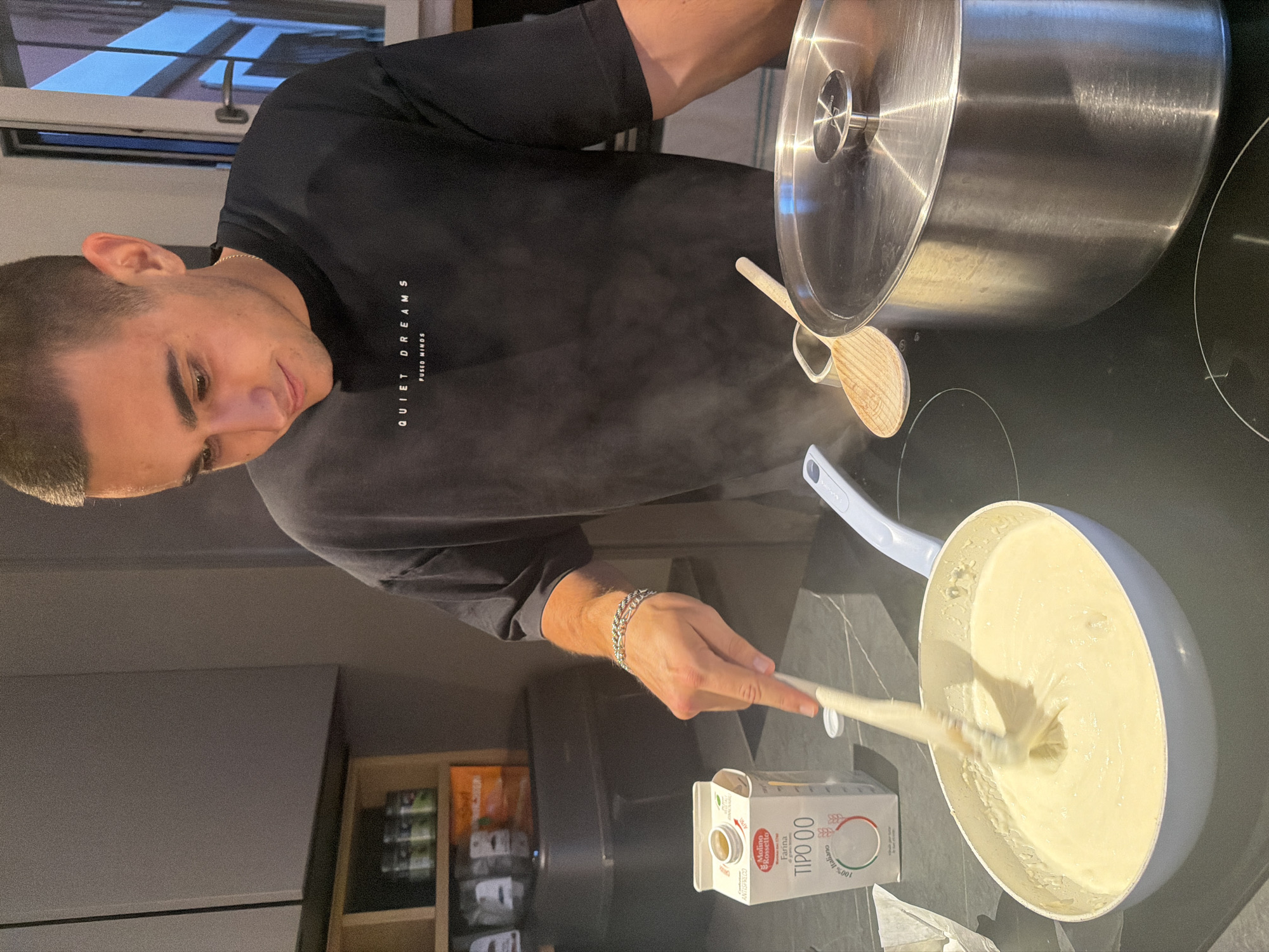 A young man with short dark hair is standing in a modern kitchen, stirring a creamy mixture in a light blue frying pan with a wooden spatula. He is wearing a black t-shirt with "QUIET DREAMS FUSED MINDS" printed on it. Next to the pan is a carton of "Molino Rossetto Farina TIPO 00 100% Italiano." A large stainless steel pot with a lid is to his right, and steam is rising from the pan. The kitchen has grey cabinets and a dark countertop. The background shows shelves with jars and containers. The overall scene depicts a man cooking in a home environment. The image does not provide any information to ascertain the location in Milan, Italy.