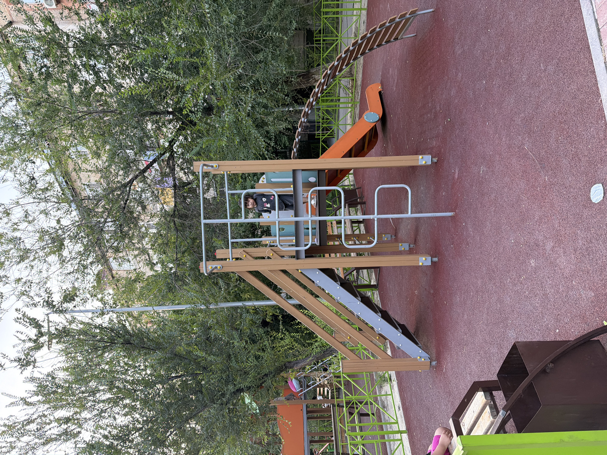 The image shows a children's playground in Shymkent, Kazakhstan. The playground features a multi-level wooden and metal structure with slides, stairs, and a climbing wall. A young child, appearing to be a girl with dark hair wearing a dark shirt, stands on the upper level of the playground. In the foreground, a section of a bench and a metal bin are visible. The playground has a red rubberized safety surface. Green metal fencing surrounds part of the playground. Trees with green foliage are present, and a multi-story building is visible in the background. The lighting suggests it is daytime.