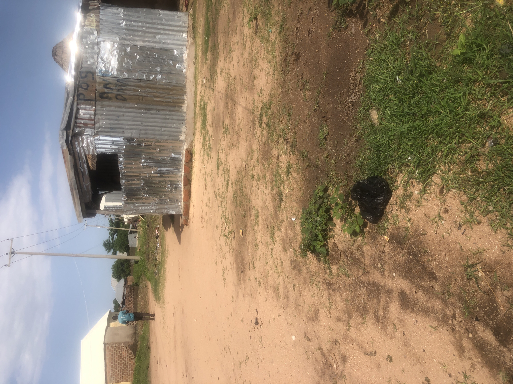 The image depicts an outdoor scene in Jalingo, Nigeria. In the foreground, a dirt path dominates the frame, with patches of dry grass and some darker, damp soil. A black plastic bag of refuse is visible on the right side of the path, near a clump of green grass.
Further into the image, a corrugated metal structure, possibly a small shop or dwelling, stands to the right. It has a thatched roof section at the rear. Graffiti is visible on the metal walls. To the left, a building with a white roof and brick construction is present. A lone individual, appearing to be a young person wearing a blue shirt and dark pants, walks away from the camera towards the building on the left.
In the background, more structures and vegetation are visible. A utility pole with overhead wires stands prominently in the middle ground. The sky is bright blue with some wispy clouds. The overall impression is of a somewhat unkempt, rural or semi-urban environment.