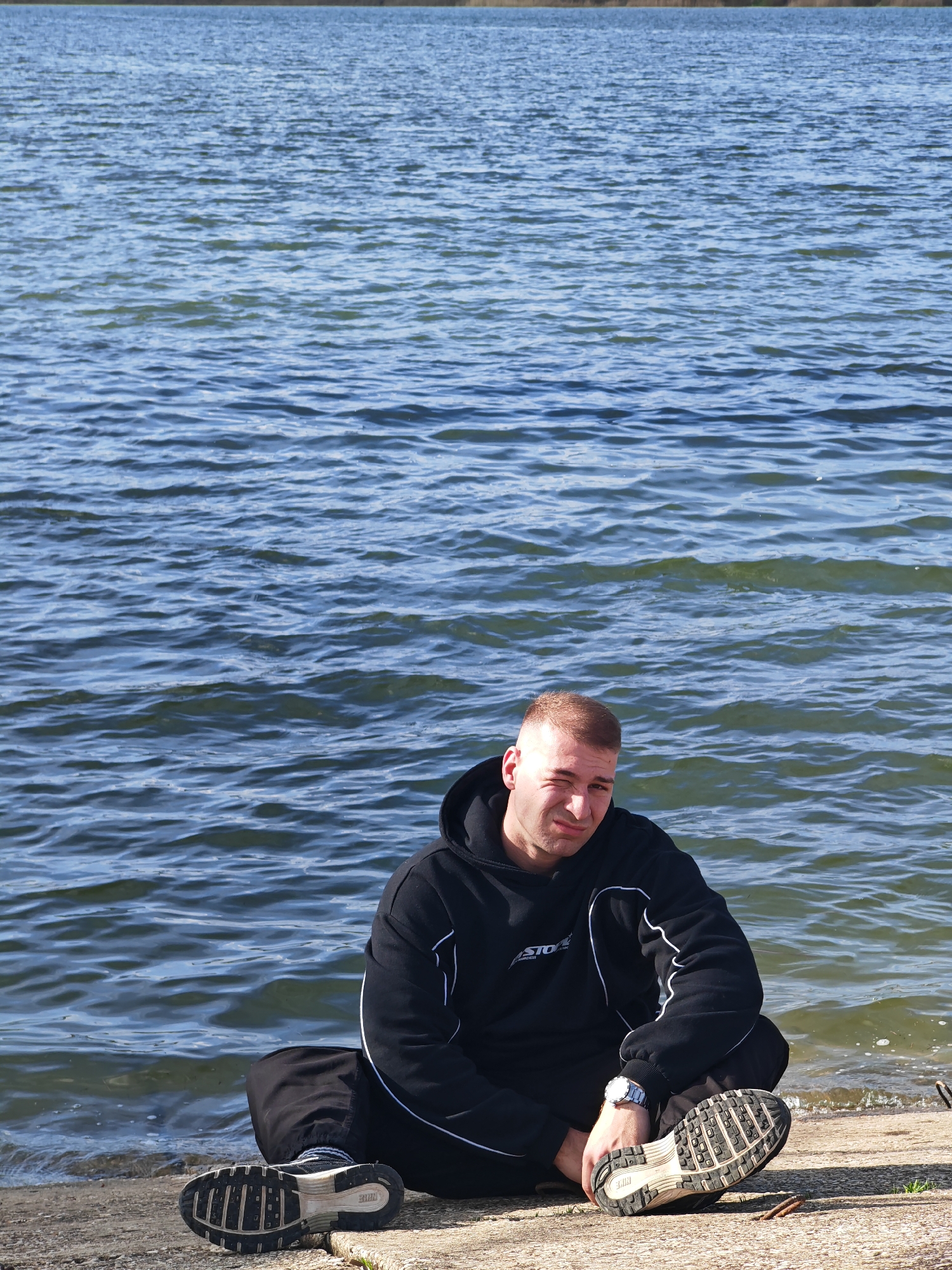 A young man sits cross-legged on a concrete ledge, with a body of water behind him. The man wears a black hooded sweatshirt with white piping along the sleeves and a watch on his left wrist. His shoes are off and placed sole-up next to him on the ledge. The water behind him is a bluish-green with small waves. The scene is likely located on the shore of a lake or reservoir in the vicinity of Shumen, Bulgaria, given the context.