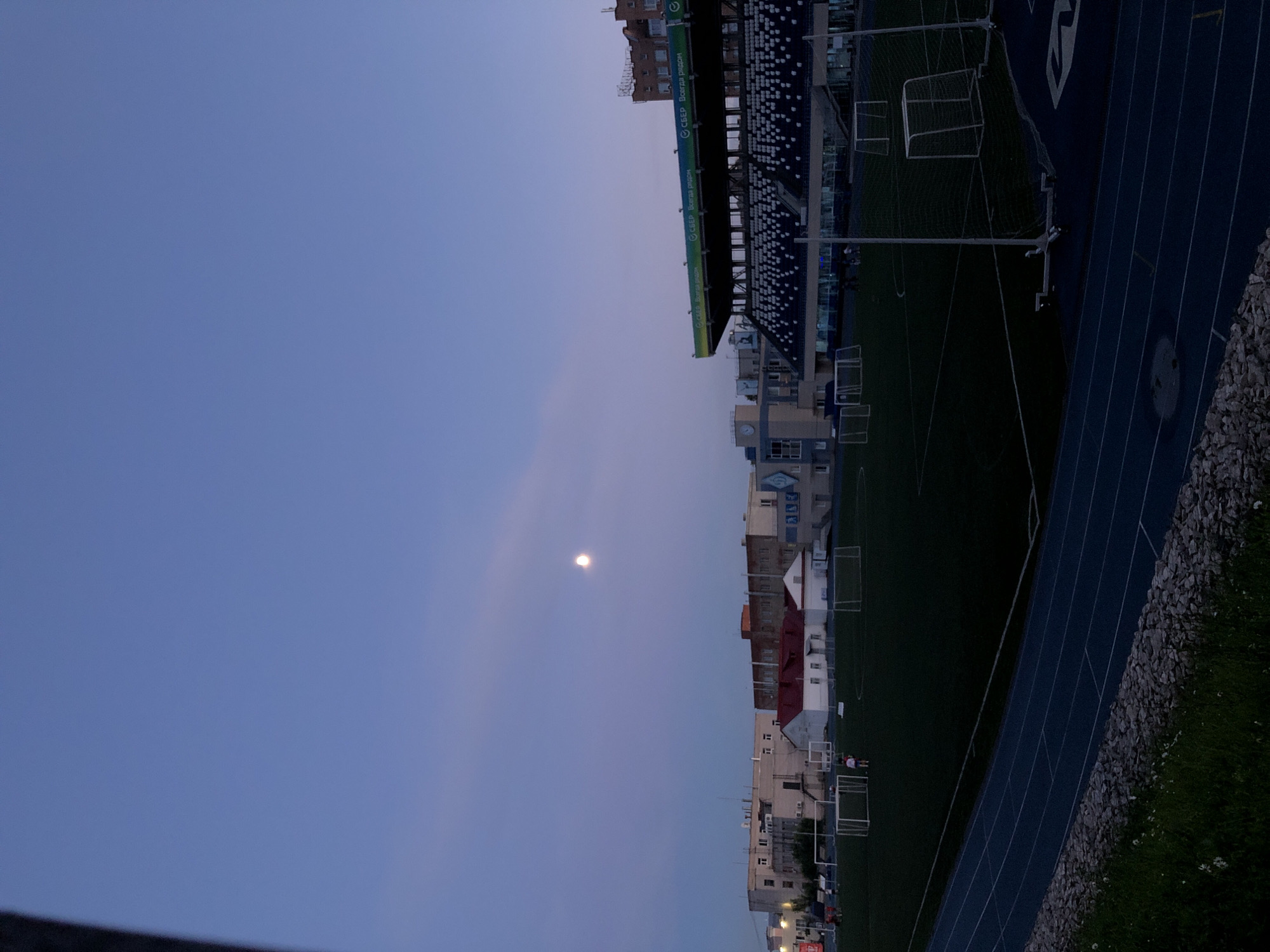 This is a photograph taken in Izhevsk, Russia, featuring an outdoor sports complex at dusk. The scene is dominated by a sports stadium with dark blue seating and a green artificial turf field, marked with white lines and soccer goals. A blue running track runs along the side of the field. Beyond the stadium, a line of buildings, some with red roofs, is visible. The sky is a pale blue, suggesting the transition from day to night, and the moon is clearly visible as a bright orb in the sky.
There are no clear indications of people engaging in any specific activity, though a few figures might be present in the distance near the goals. The overall atmosphere is tranquil, with the twilight setting and the prominent moon creating a calm and serene mood. The image is oriented vertically, meaning the scene is presented sideways.
Text is visible on the stadium's facade, appearing as a green strip with white lettering. Partially legible, it seems to say "СБЕР. Всегда рядом" which translates to "Sberbank. Always close," indicating a sponsorship or advertising placement. The weather appears clear, with no clouds obscuring the sky or the moon. The location is identifiable as Izhevsk, Russia.