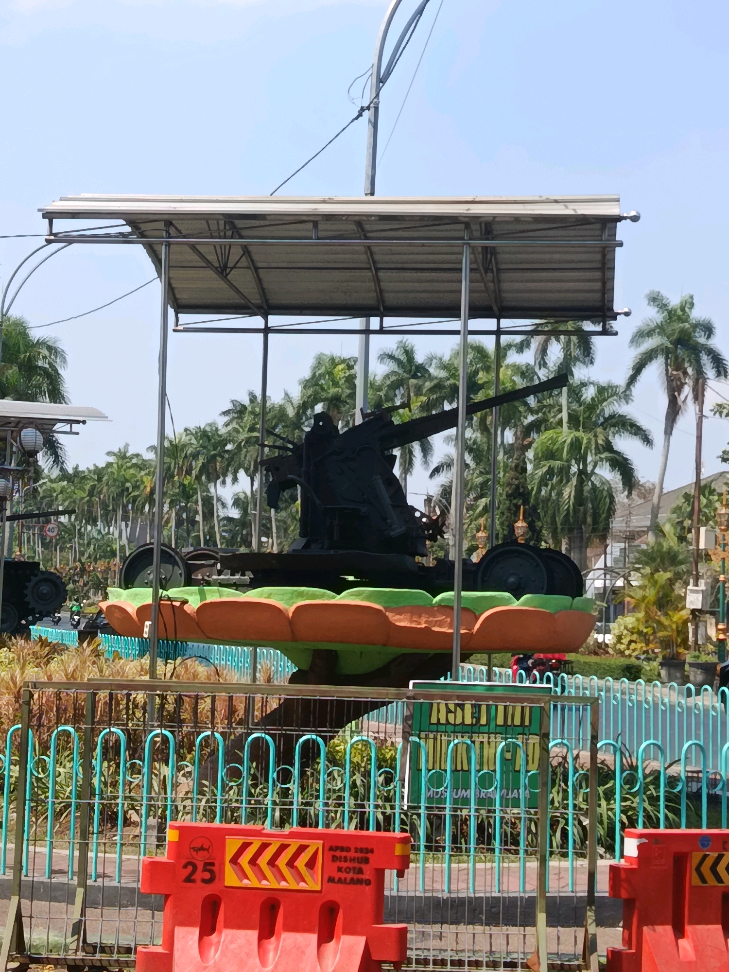 In Malang, Indonesia, a large, black anti-aircraft gun is on display. The gun is mounted on a base that resembles a flower with orange and green petals. It is positioned under a metal canopy supported by poles. In the background, tall palm trees and buildings are visible. A teal metal fence surrounds the display, and in the foreground, red traffic barriers are present. One barrier has the text "APBD 2024 DISHUB KOTA MALANG" and the number "25" printed on it.
