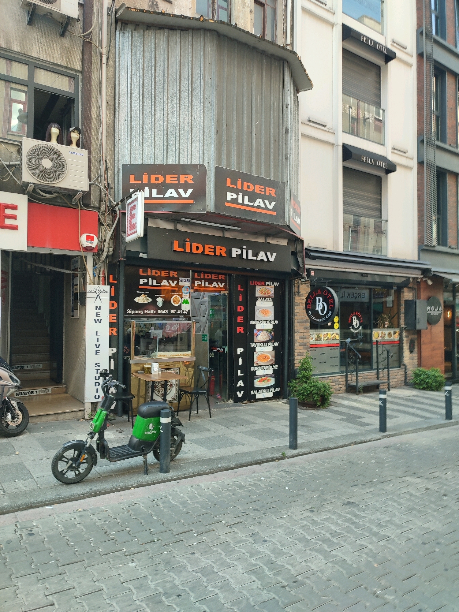 The image depicts a street scene in Kadıköy, Turkey, focusing on the storefronts of several businesses. The primary subject is a restaurant called "LİDER PİLAV" with prominent orange signage featuring the name in white lettering. Next to it, the "BELLA OTEL" is visible, a hotel with white facade and black awnings. In front of the restaurant, an electric scooter, painted green with a black seat and a white logo, is parked on the sidewalk. The street is paved with cobblestones, and there are bollards along the edge of the sidewalk. The architecture suggests a bustling urban environment. To the left of the "LİDER PİLAV" is another establishment with a red facade and a window display showing what appear to be mannequins. The overall impression is that of a commercial street with various retail and hospitality businesses.