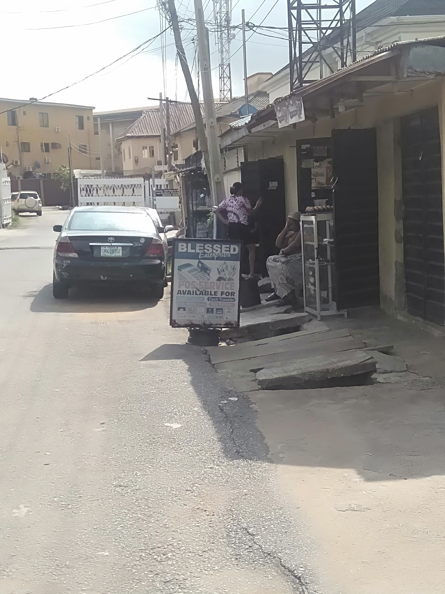 The scene is a street in Shangisha, Nigeria. A black sedan is parked on the left side of the road facing away from the viewer. Next to the car, a sign reads "BLESSED Enterprise POS SERVICE AVAILABLE FOR". Two people are near the entrance of a building on the right. One is standing and wearing a pink and white shirt with dark shorts. The other is seated, wearing a patterned dress and a head covering. A metal display case is next to the seated person. The building has a partially open black door. There is an uneven concrete area in front of the building. The street is paved and appears to be in moderate condition. Buildings are visible on both sides of the street. Overhead power lines and poles are present.