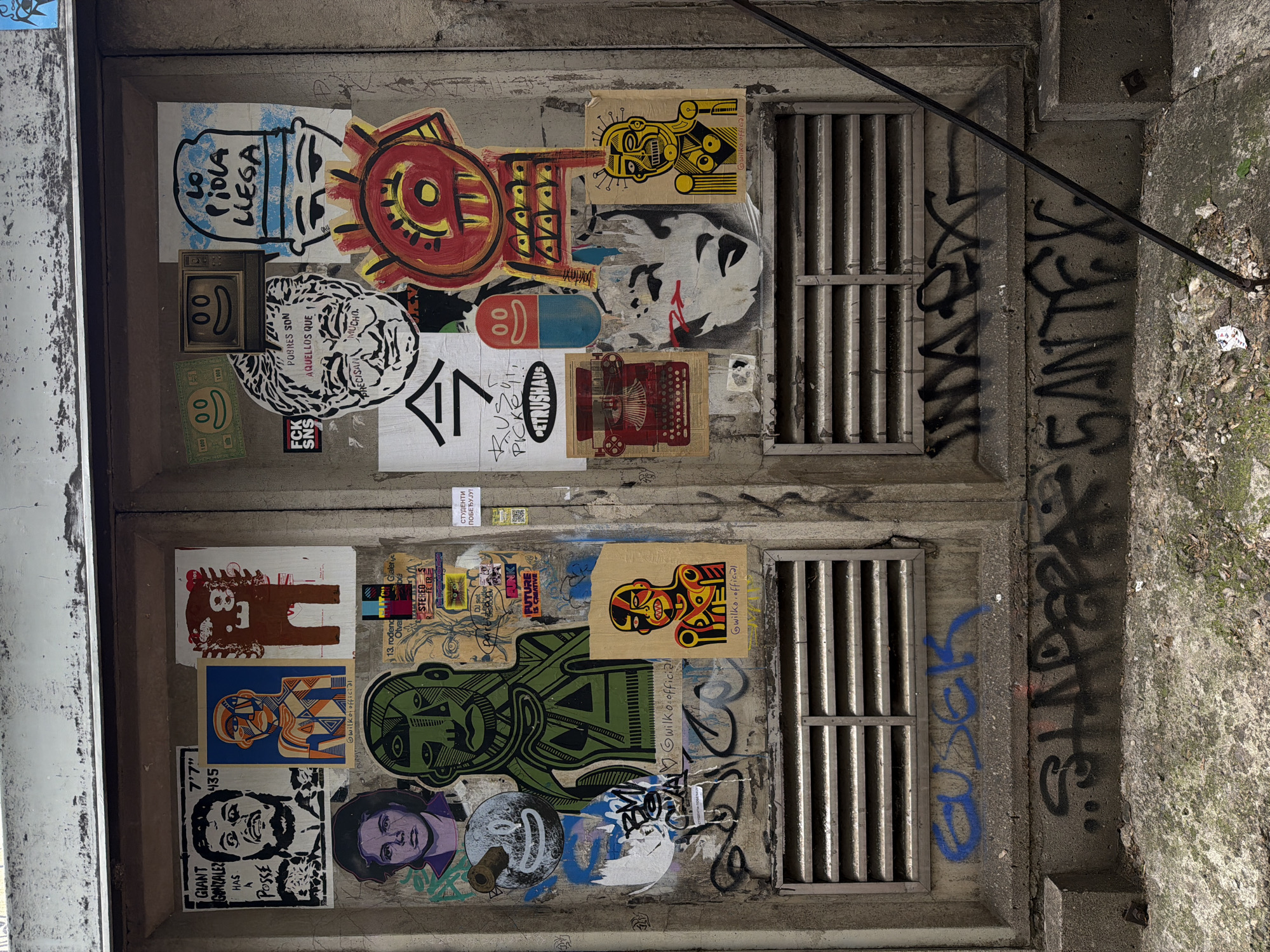 The image shows a textured concrete structure, likely a basement access point, covered in graffiti and stickers. Two vertically oriented metal grates are set into the structure, one above the other. The left side of the structure, appearing to be doors or panels, is heavily decorated with a variety of stickers and artwork. These include stylized portraits, geometric designs, and text in both English and possibly Serbian or Spanish. Specific stickers show faces, patterns, and phrases like "LA PIOLA LLEGA," "POBRES SON AQUELLOS QUE PRECISAN MUCHO," "GIANT GONZALEZ HAS A POSSE," and what seems to be social media handles (@wilko.official). Graffiti includes the name "SHAPEPA SANTEX." The concrete has varying levels of wear and discoloration. The location is in Belgrade, Serbia, evidenced by the Serbian text "СТУДЕНТИ ПОБЕЂУЈУ!".