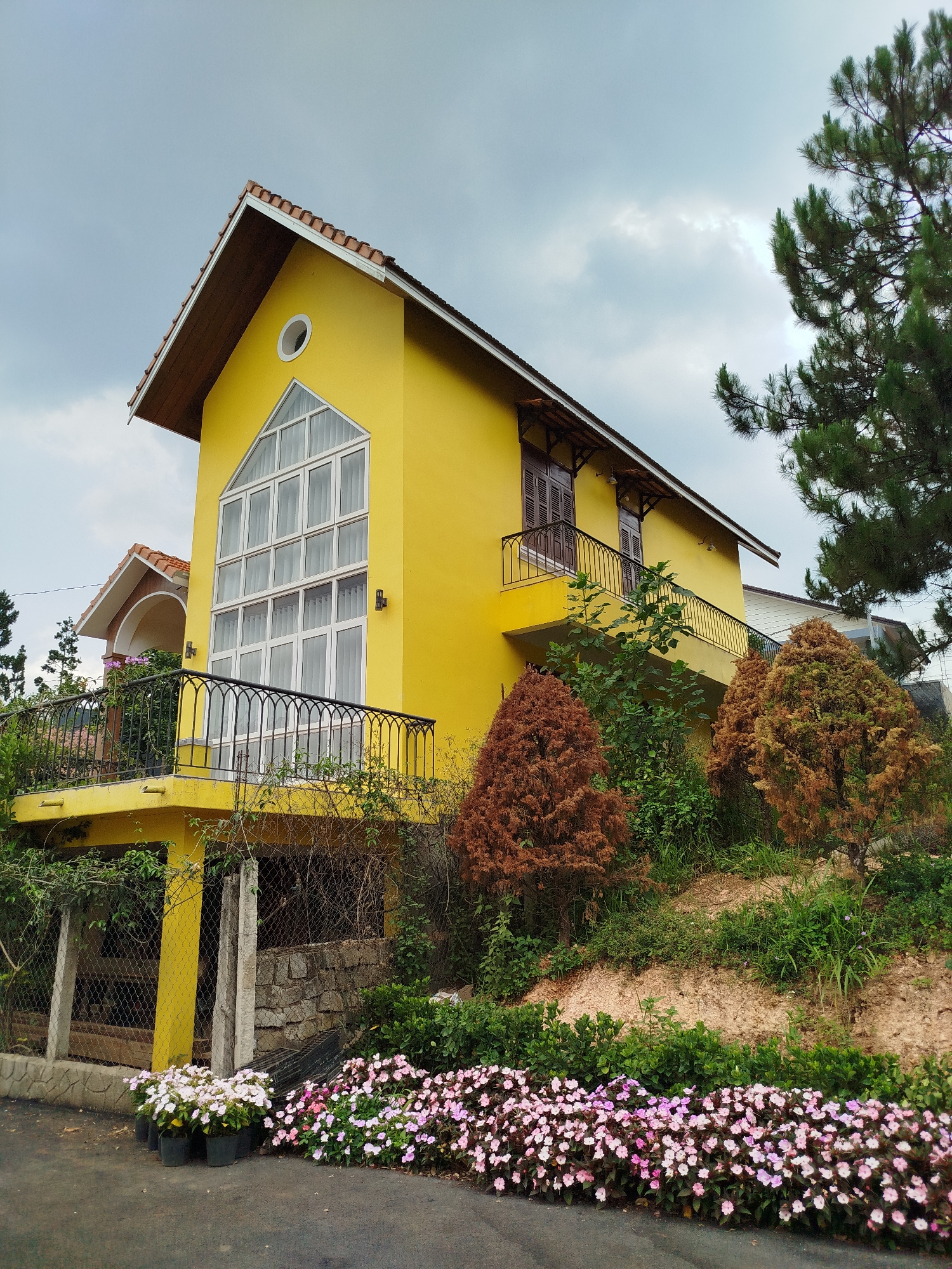 The image shows a two-story yellow building with a brown roof, likely a house, located in Hiep Thanh, Vietnam. The house has a large window with a triangular top and multiple panes, a balcony with dark railings, and brown shutters on the upper floor. The building sits on a small embankment covered in greenery and reddish-brown bushes. A row of pink and white flowers lines the base of the embankment along the road. A chain-link fence runs along the left side of the building. A large green pine tree is visible to the right, partially obscuring the house. The sky is overcast.