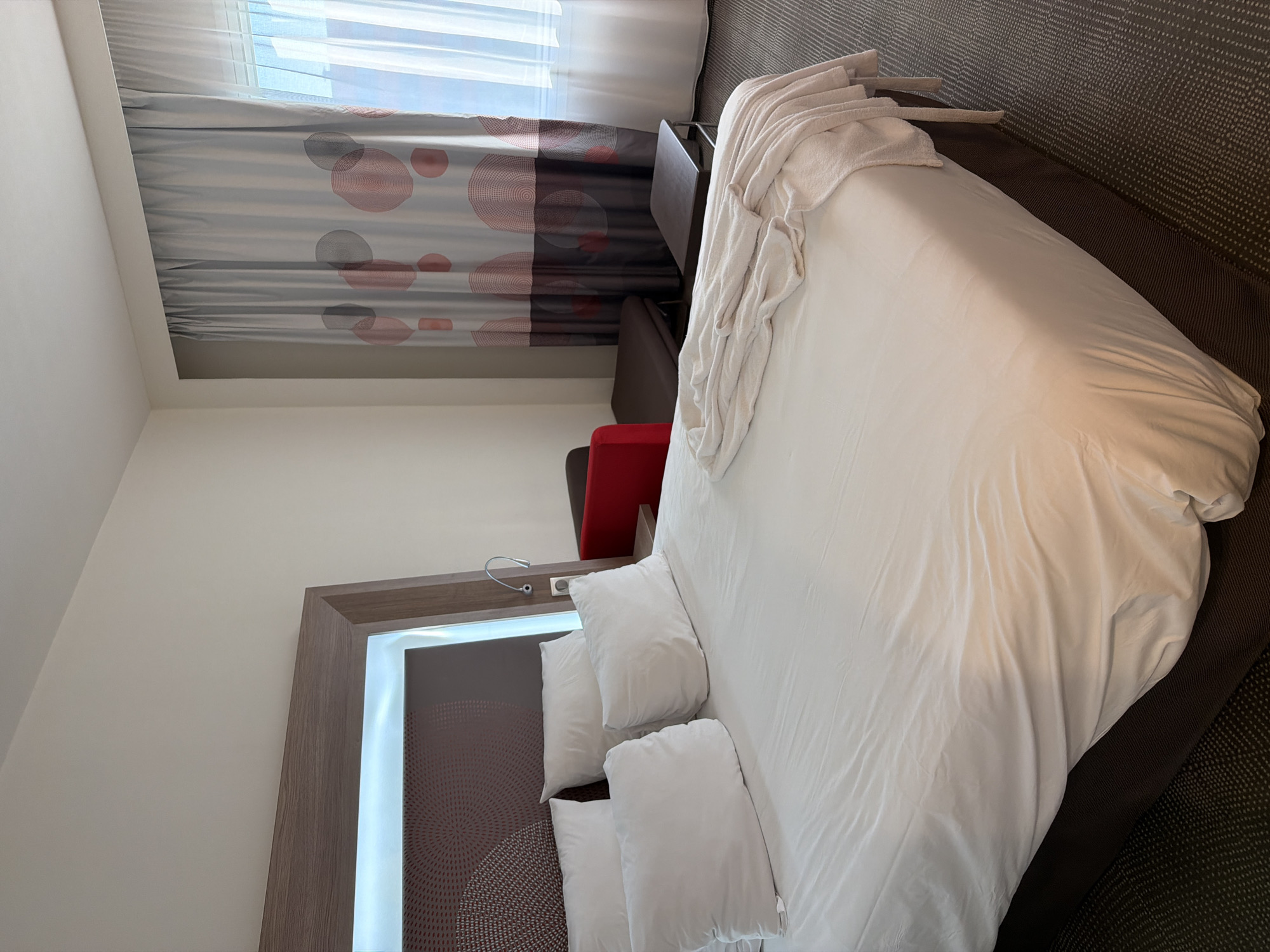 A hotel room in Khimki, Russia, features a bed with white linens and several white pillows. A folded white robe or blanket lies on top of the duvet. The headboard is brown with a circular pattern and is framed by a wooden structure with integrated lighting. To the right of the bed, a red sofa-like seating arrangement is partially visible, along with a small dark brown table. The room has patterned grey carpet and a window covered by two layers of curtains: a patterned grey and red fabric overlaying sheer white panels.