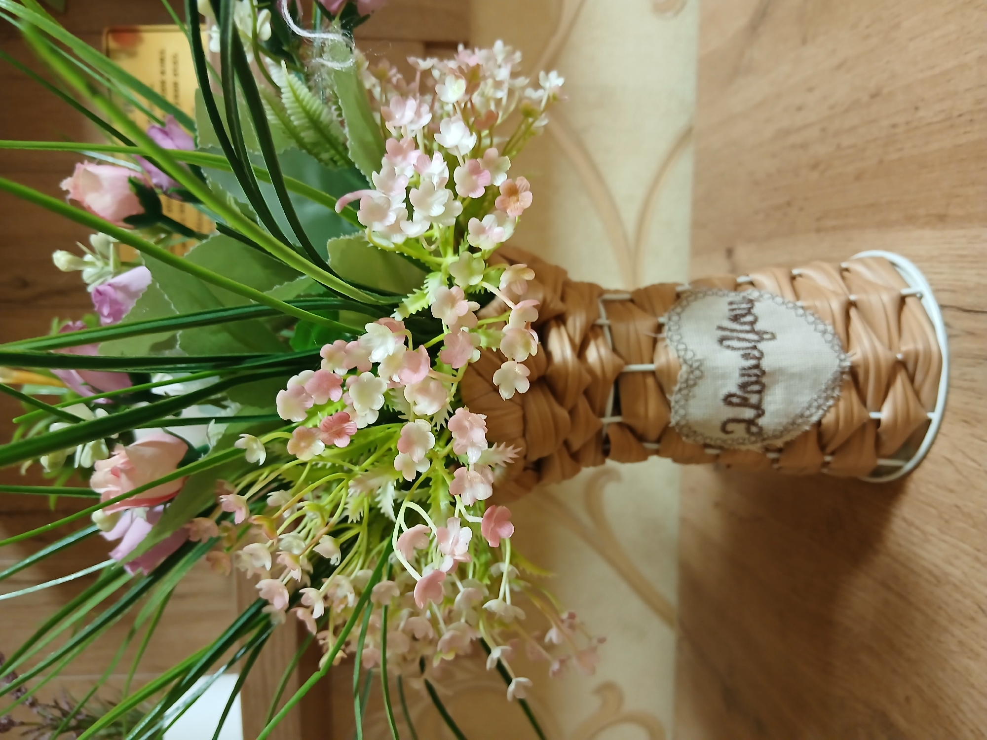 A close-up, eye-level shot captures a decorative vase filled with artificial flowers. The vase, made of woven brown material, features a small heart-shaped patch with the embroidered phrase "I love you." The flowers are a mix of light pink roses and smaller, delicate pink and white blossoms, interspersed with green foliage and grass-like stems. The vase rests on a wooden surface, possibly a table, with a patterned wallpaper visible in the background. The lighting is even, highlighting the textures and colors of the arrangement.