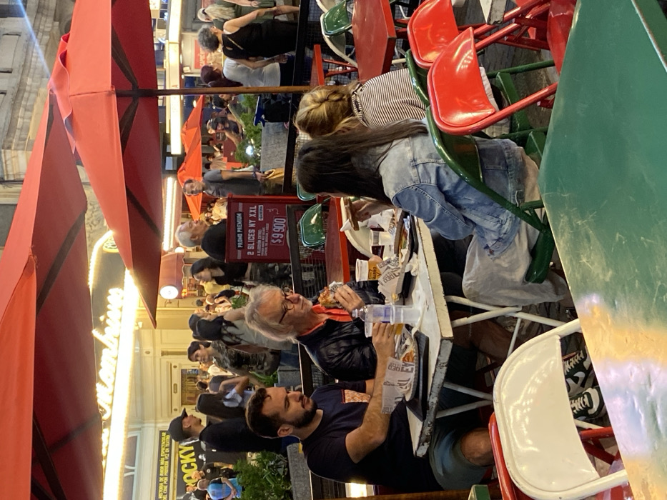 A group of people dine outdoors under red umbrellas, at tables with green and white chairs. Some individuals are eating, including a man with gray hair, while others are engaged in conversation. A sign advertises a special offer of "2 slices NY XXL" for $9,900. In the background, there is a building with illuminated signage and a crowd of people.