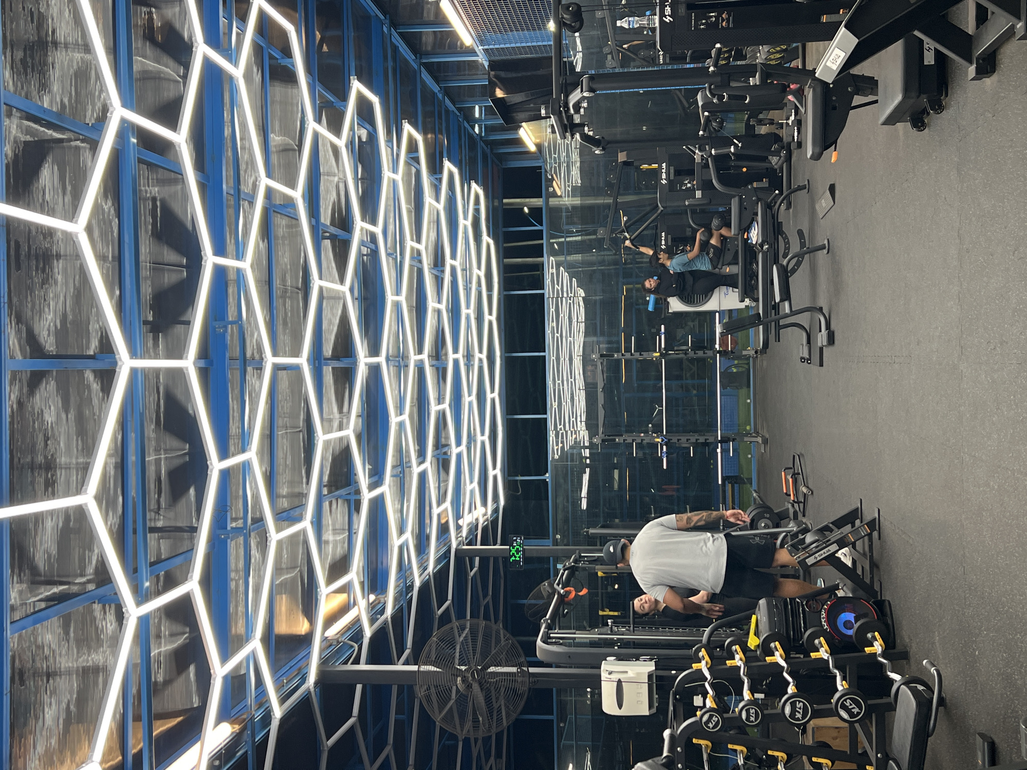 The image depicts the interior of a modern gym, likely located in Arica, Chile, given the context. The ceiling is adorned with an intricate network of hexagonal LED lights against a blue background. In the foreground, a gym rack with various weights and barbells is visible on the left. To the right of this rack, a man wearing a grey t-shirt and black shorts stands facing away from the camera, with his back to the viewer. Next to him, a woman in dark clothing stands with her hands on her hips. Further into the gym, a man in a blue t-shirt and shorts is exercising on a weight machine. Another weight machine is visible to the right, with a person partially obscured by it. A large industrial fan is mounted on a pole on the left side of the image. A digital clock displaying "7:25" is mounted on the wall in the background. The floor is covered with black rubber mats. The overall environment suggests a functional and well-equipped fitness facility.