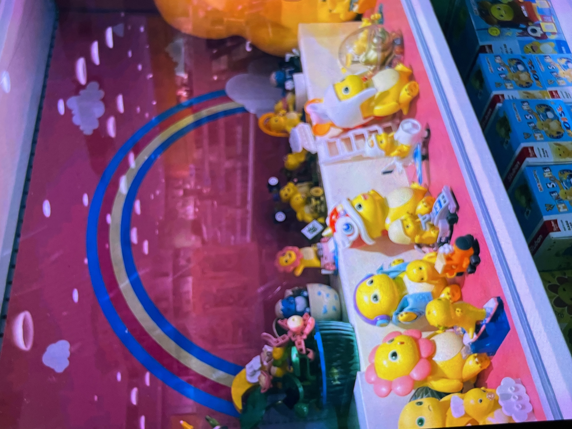 The image depicts a brightly lit display shelf filled with various toy figurines and boxes. The backdrop features a pink wall with a painted rainbow and clouds, suggesting a playful or children's themed setting. The toys are predominantly yellow, round-bodied figures with smiling faces, some wearing hats, headphones, or animal costumes. There are also smaller toy vehicles, such as a miniature orange train and a blue skateboard. The toys are arranged on a white shelf, with some placed in front of boxes labeled with Chinese characters and the word "M AS S Ms." The overall impression is of a toy store display. There is no information in the image to suggest it is located in Cadek, Indonesia.