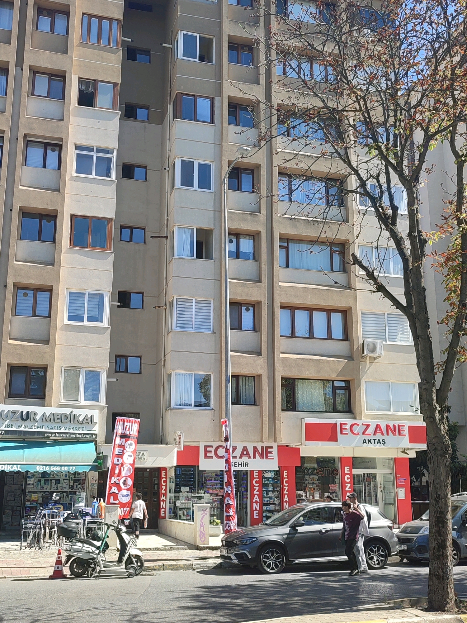 The image shows a street view in Kadıköy, Turkey. A multi-story apartment building with numerous windows is the dominant feature. In front of the building, there are several storefronts, including "UZUR MEDİKAL" and "ECZANE AKTAŞ" (pharmacy). A white scooter is parked on the sidewalk, and two people are walking in front of the pharmacy. A grey car is parked in the street, and another car is partially visible. A tree with bare branches and some autumn foliage is located on the right side of the image. The sky is clear and the sun is casting shadows.