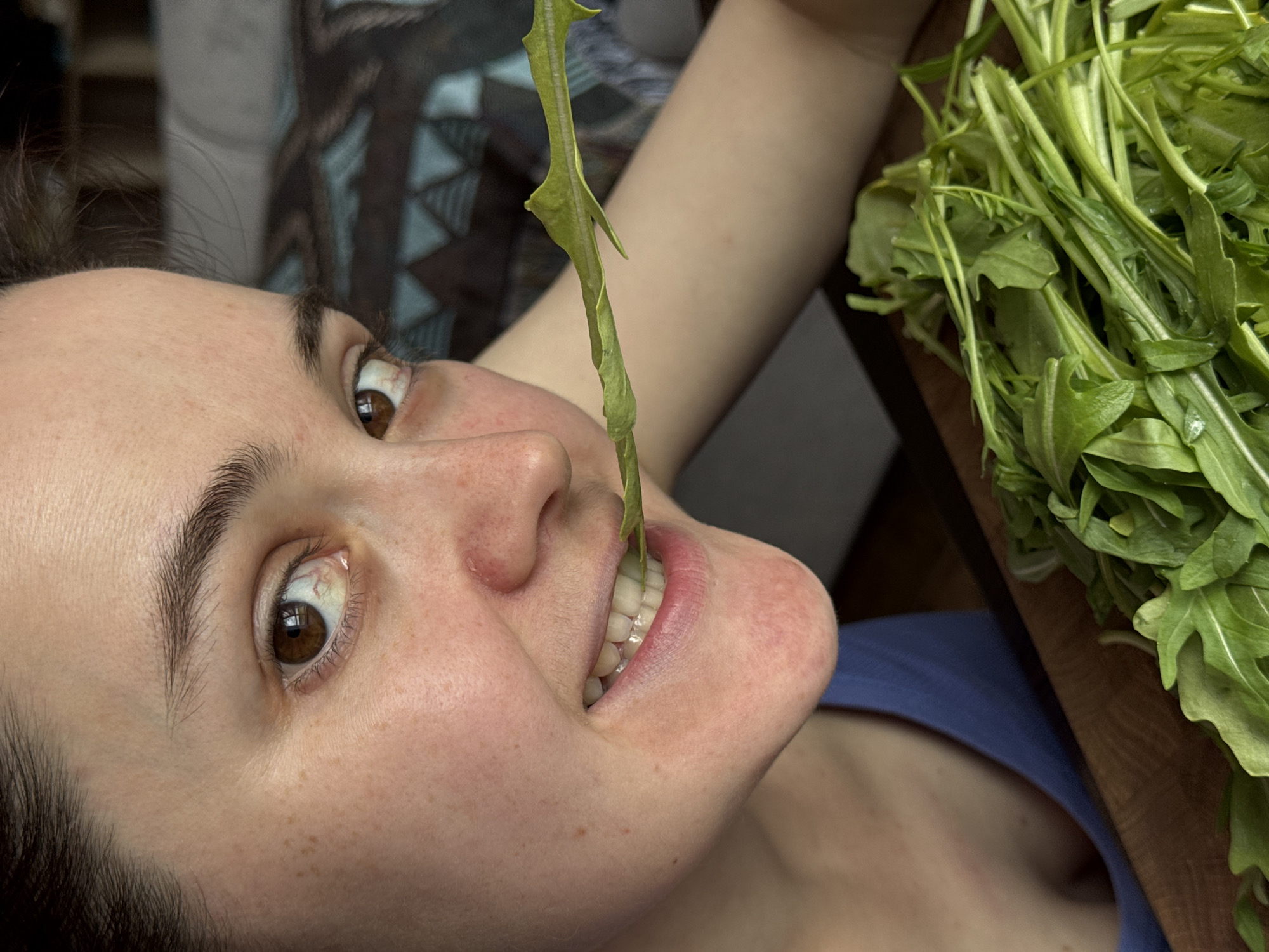 A woman is angled in the frame with a stem of leafy greens in her mouth. The greens, presumably arugula based on the pile to the right of the frame, are bright green. The woman has brown eyes and her hair is dark. Her skin has freckles. She wears a blue shirt. The background includes a decorative patterned textile and a dark wooden surface. No information in the image suggests this photo was taken in Izhevsk, Russia.