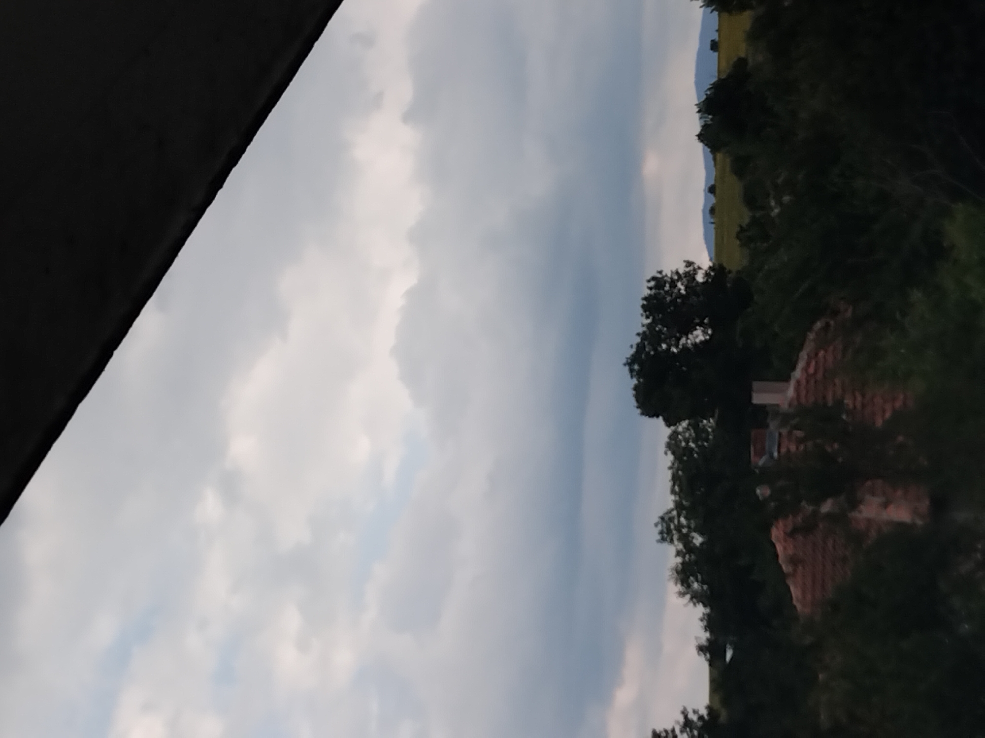 The image captures a view from a higher vantage point, looking out over a landscape dominated by a cloudy sky. In the foreground, a dark, angled structure, possibly a roof overhang or beam, partially obscures the upper portion of the frame. Below this, a dense canopy of dark green trees stretches across the midground, interspersed with glimpses of a reddish-tiled roof of a building. One building shows a visible chimney stack. Beyond the trees, rolling hills are visible under the cloudy sky, with hints of yellow in the fields suggesting agricultural land. The overall impression is one of a natural or semi-rural setting, possibly on the outskirts of a town or village, given the presence of the building. The location is identified as Plastina, Bulgaria.