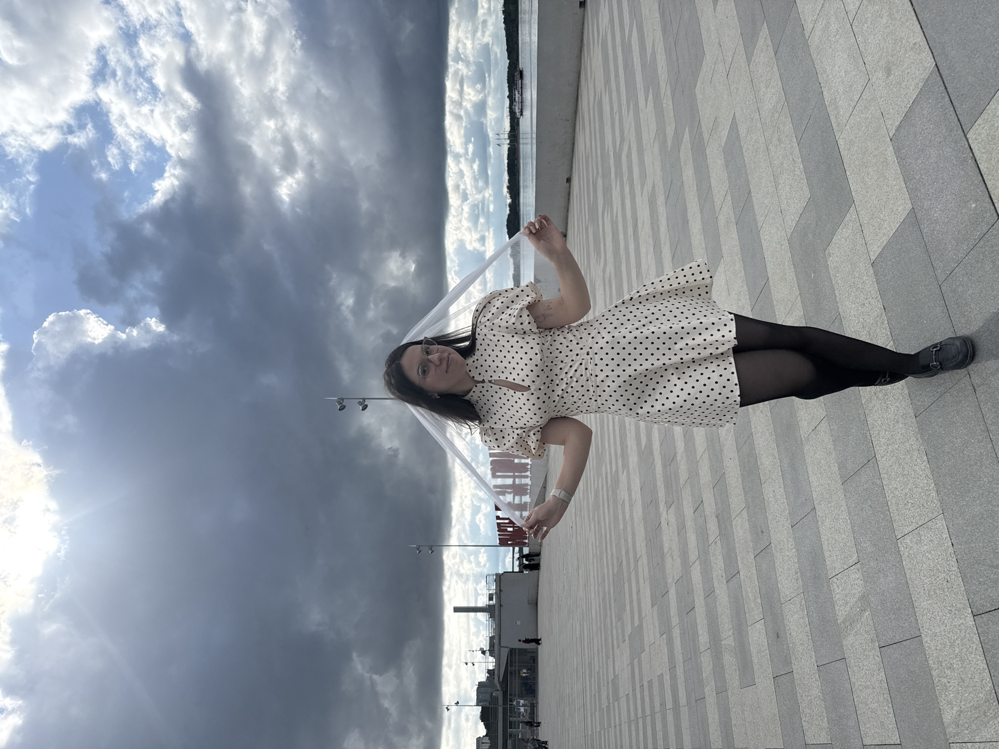 A young woman stands centrally on a paved promenade under a dramatic, cloudy sky with intermittent sunlight. She wears a cream-colored, polka-dotted dress with short puffed sleeves and a high collar with a keyhole cutout, black opaque tights, and dark loafer-style shoes. A white wristband adorns her left wrist. She holds a transparent banner-like object with both hands, extending it outwards, on which the Russian word "СЧАСТЬЕ" (Happiness) is visible in dark lettering.
To her left, the promenade extends towards several grey buildings, one resembling a terminal entrance with turnstiles, and multiple streetlights. In the mid-ground, a large red art installation spelling out "СЧАСТЬЕ НЕ ЗА ГОРАМИ" (Happiness is not far off) is partially visible. To her right, a wide body of water, identified as the Kama River, stretches into the distance, with a forested bank beyond. A white passenger boat and a sailboat are visible on the river. The scene is set on the Kama River embankment in Perm, Russia, identifiable by the prominent "СЧАСТЬЕ НЕ ЗА ГОРАМИ" art object. The sky is predominantly overcast with dark, heavy clouds, though a bright patch of sun breaks through on the upper left, and areas of blue sky are visible through cloud breaks on the right. The promenade is paved with rectangular grey and lighter grey tiles laid in an alternating pattern.