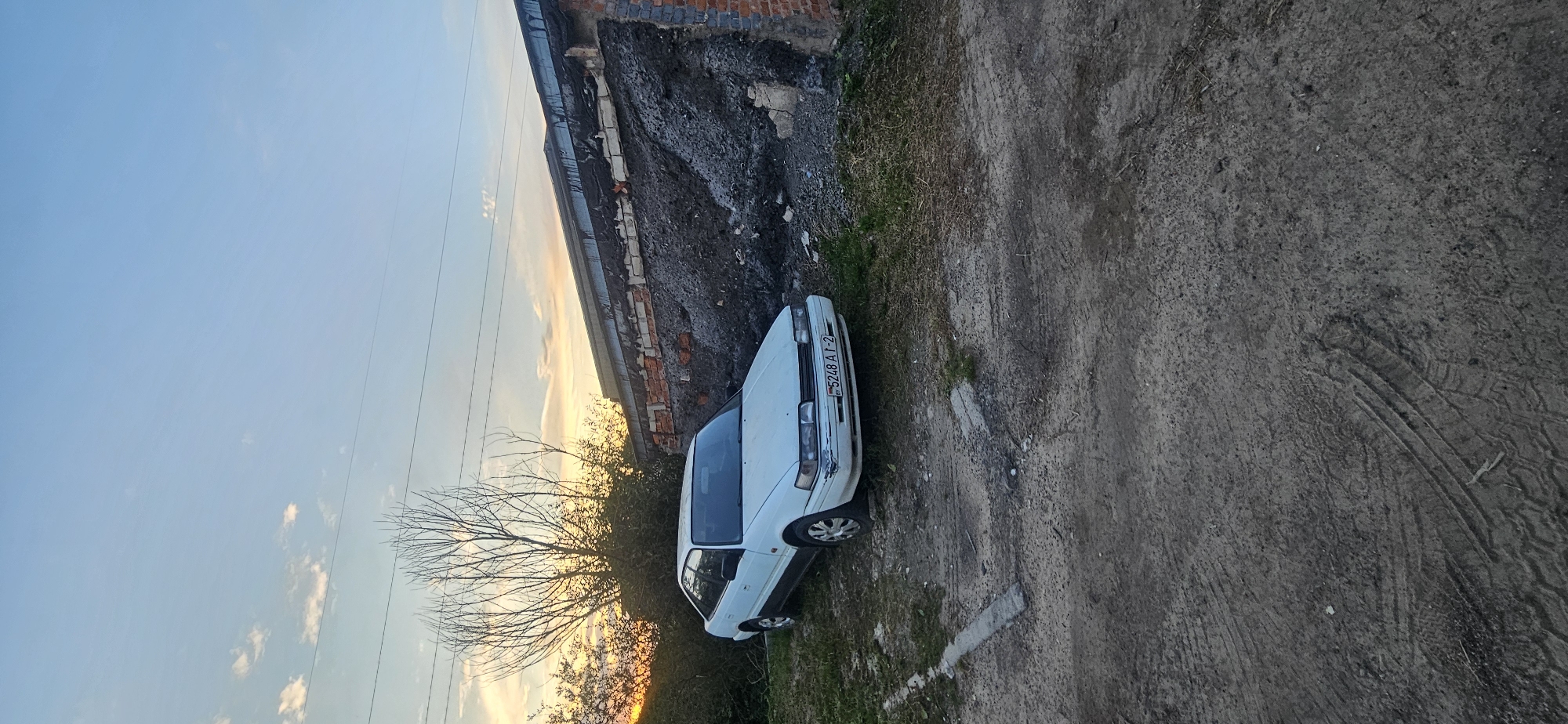 In the foreground, a dirt and gravel surface exhibits distinct tire tracks. A white four-door sedan, identified by the license plate "BY 5248 AT-2", is parked on a grassy area next to a damaged brick and concrete structure. This structure appears to be a partially collapsed garage or outbuilding, with exposed brickwork and debris. Behind the car and to the left, a bare-branched tree and overgrown bushes are silhouetted against a sky with soft clouds and a sunset glow. The environment suggests a rural or semi-abandoned setting within the Mazalawski Sel'ski Saveet, Belarus.