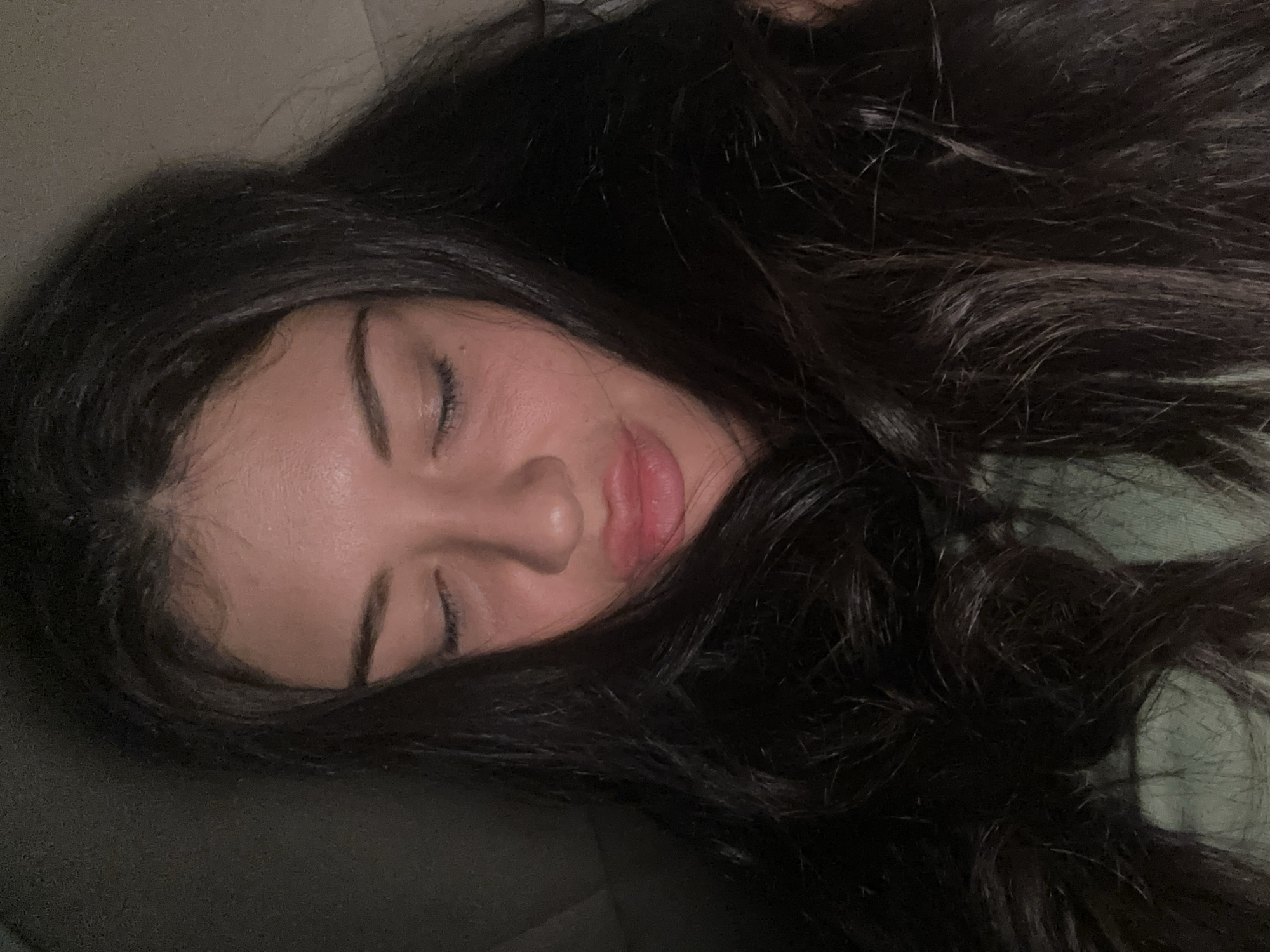 The image captures a close-up, high-angle shot of a young woman resting indoors, likely within a home environment in Khimki, Russia. The primary subject is the woman, who appears to be lying down on her side, with her face visible in profile. She has long, dark, wavy hair that cascades around her head and upper body. Her eyes are closed, and her facial expression is serene and relaxed, with her lips slightly parted in a soft, natural position.
Her skin tone is fair, and faint shadows suggest soft, indirect lighting, indicating it might be nighttime or a dimly lit room. No distinct light sources are visible. She is wearing a light green garment, possibly a shirt or blanket, which is visible on her right side. A muted, lighter-colored surface, possibly a pillow or headrest, is behind her head. The overall mood is peaceful and calm. There is no visible text in the image. The setting appears comfortable and private, consistent with a bedroom or living room.