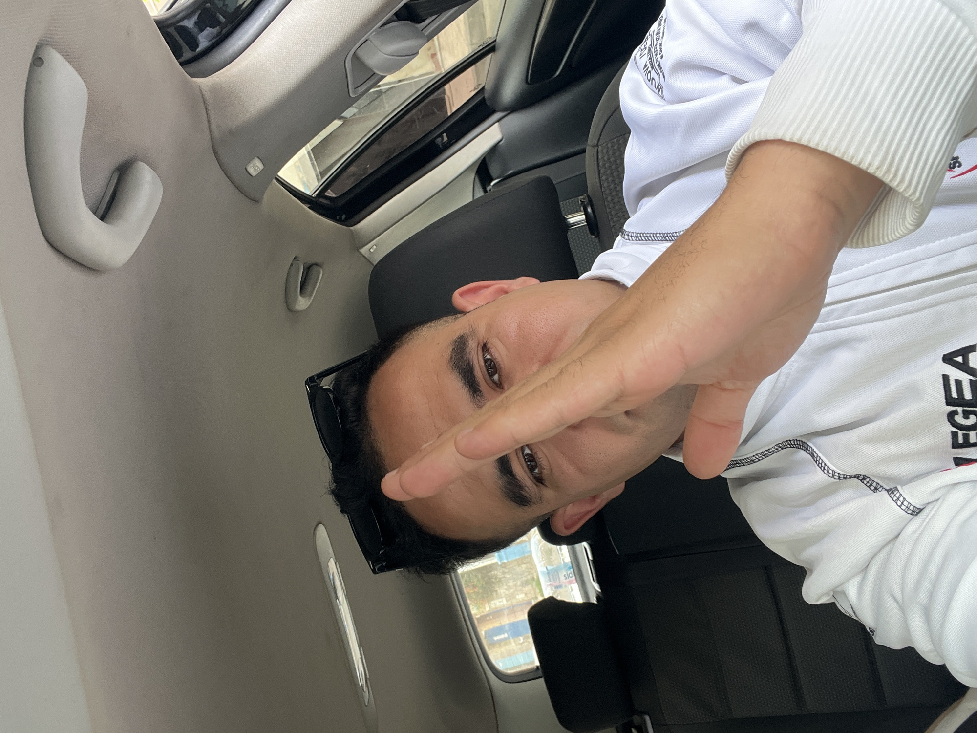 A man is depicted inside a car, posing for a photo, likely a selfie. The image is rotated counter-clockwise, showing the man's head to the left and his body extending to the right. He has dark hair and brown eyes, looking directly at the camera. Black sunglasses are pushed up onto his head. His right hand is partially covering his face, with fingers spread in a V-like gesture over his eyes.
He is wearing a white long-sleeved shirt with ribbed cuffs. The brand name "LEGEA" is visible in black text on his right sleeve, near the forearm. Partially visible text, possibly "KUOWIA," can be seen on the left shoulder area of his shirt.
The setting is the interior of a car, characterized by a light gray headliner and pillars. A black car seat headrest is visible behind him. Car features include a sunroof overhead and light gray grab handles on the side pillars. An "AIRBAG" indicator is visible on one of the pillars. Through the car's side window, an exterior scene of buildings with light blue elements is visible, suggesting an urban or suburban environment during daytime. This scene is consistent with a location such as Martil, Morocco.