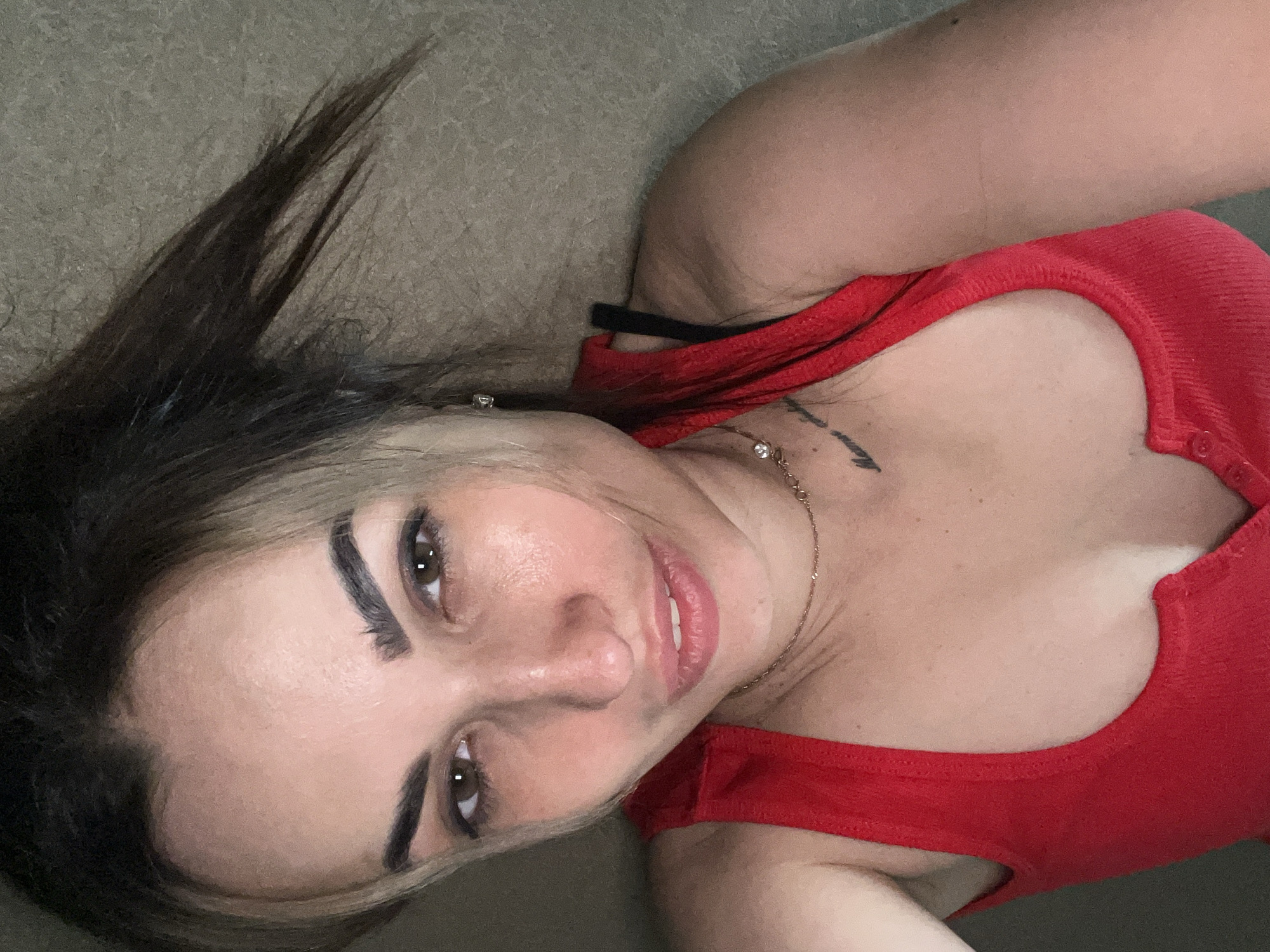 A diagonal, close-up portrait features a person, likely a woman, lying down and looking directly at the camera with a subtle smile. She has dark hair, styled with some lighter streaks near her face, which appears somewhat disheveled and spreads upwards from her head. Her face shows clear skin, prominent dark eyebrows, and brown eyes. Her lips are slightly parted, revealing a glimpse of her teeth. There is a slight sheen on her forehead and nose.
She is wearing a ribbed, red tank top or camisole, and a black bra strap is visible on her right shoulder. A delicate gold-colored necklace with small pendants, one appearing to be a clear stone, adorns her neck and chest. A second, larger pendant with multiple clear stones is visible lower on her chest. A small, dark tattoo with illegible text is visible on her left collarbone area.
The background is a uniform, textured surface in a light brown or greyish-brown hue, suggesting a sofa, bed, or carpet. The lighting is soft and even, highlighting her features without harsh shadows, indicating an indoor setting. No specific time of day or weather conditions are discernible, and there is no legible text other than the indistinct tattoo.