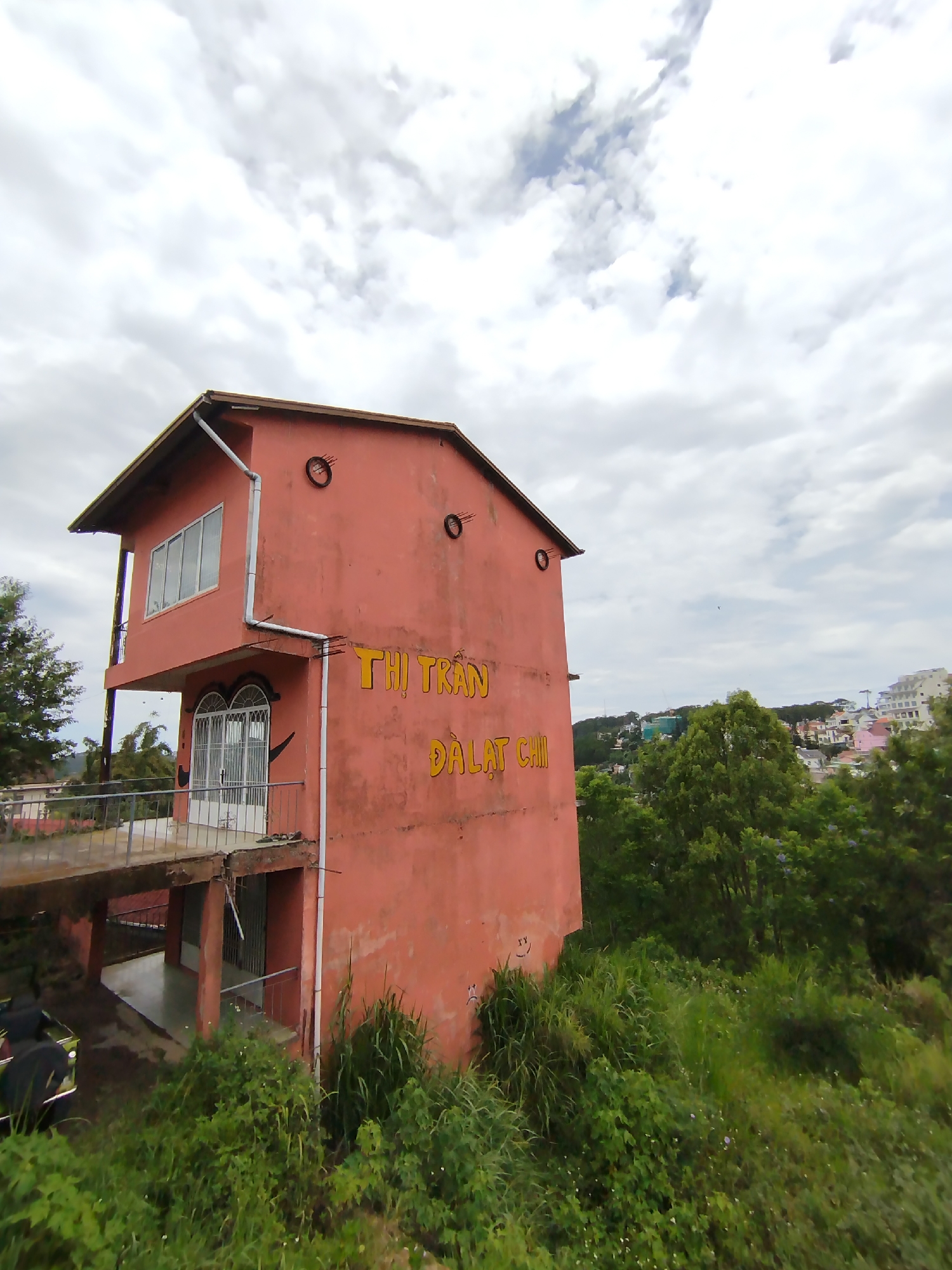 The image shows a two-story, coral-colored building in Da Lat, Vietnam. The building has a balcony on the second floor with a metal railing. The words "THI TRAN DALAT CHI" are painted on the side of the building in yellow letters. The building is surrounded by lush green vegetation. In the background, there are other buildings and trees. The sky is cloudy.